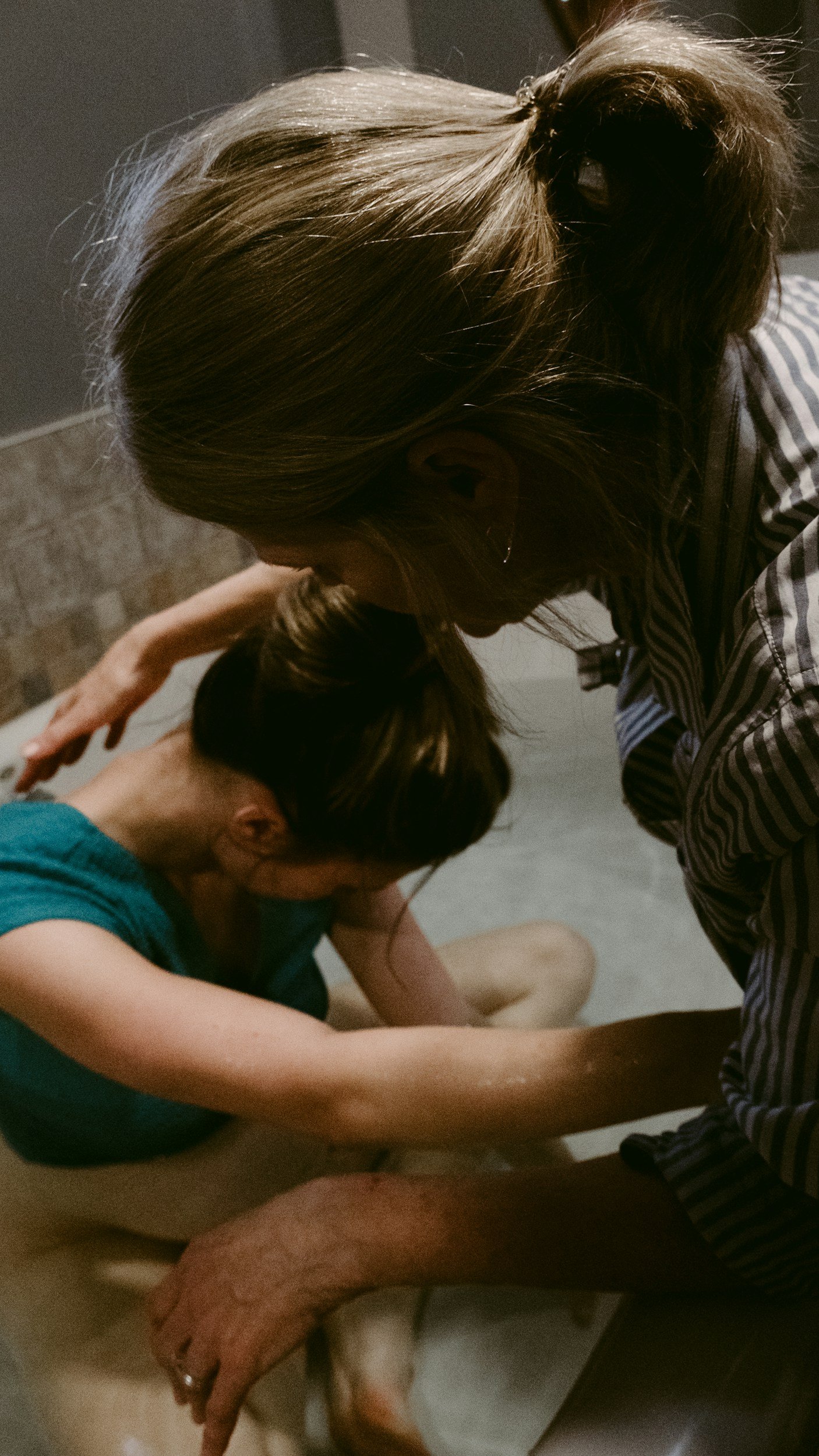 Two women are sitting on the floor, a doula in a blue shirt and the other in a striped shirt, with the striped shirt woman helping the blue shirt woman give birth support.