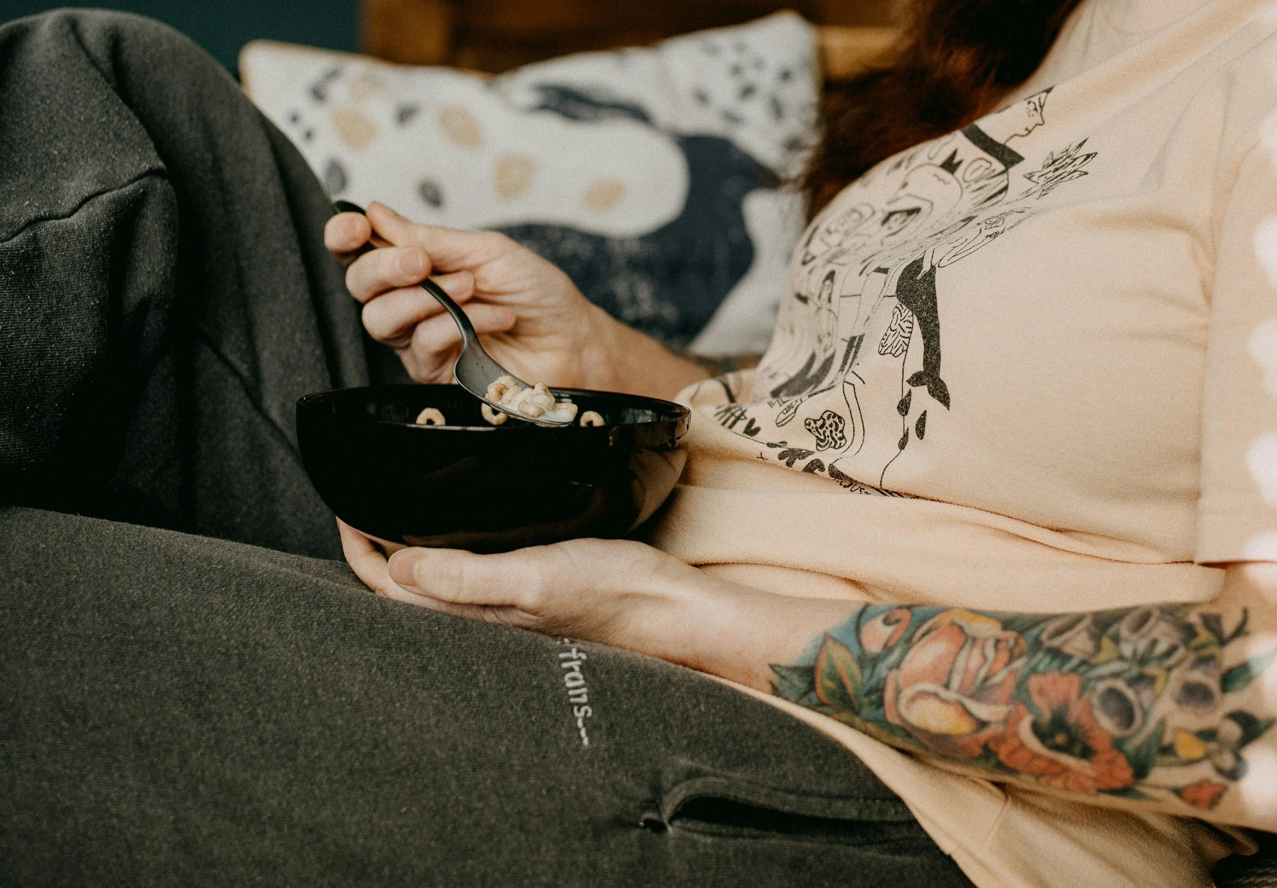 Pregnant person wearing a beige t-shirt with black comic-style illustrations, holding a black bowl with cereal, sitting next to a doula dressed in black, with a white and black patterned blanket or cloth draped over their legs.