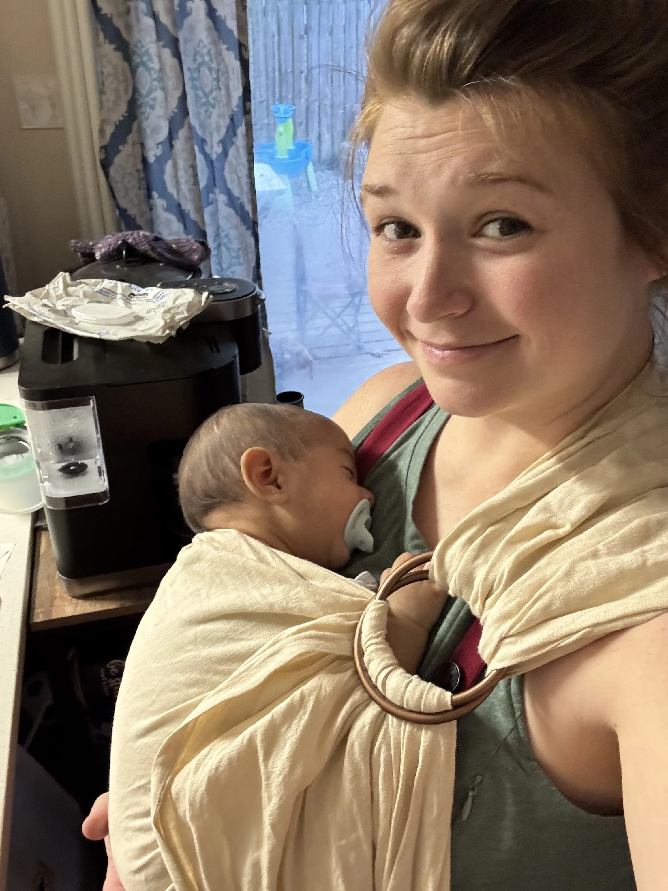 A woman is holding a sleeping baby. The woman has brown hair and is smiling slightly. The babyis resting against her chest. In the background, there is a bathroom door, a blue table with a green and blue toy, and a black appliance with a crumpled paper on top.