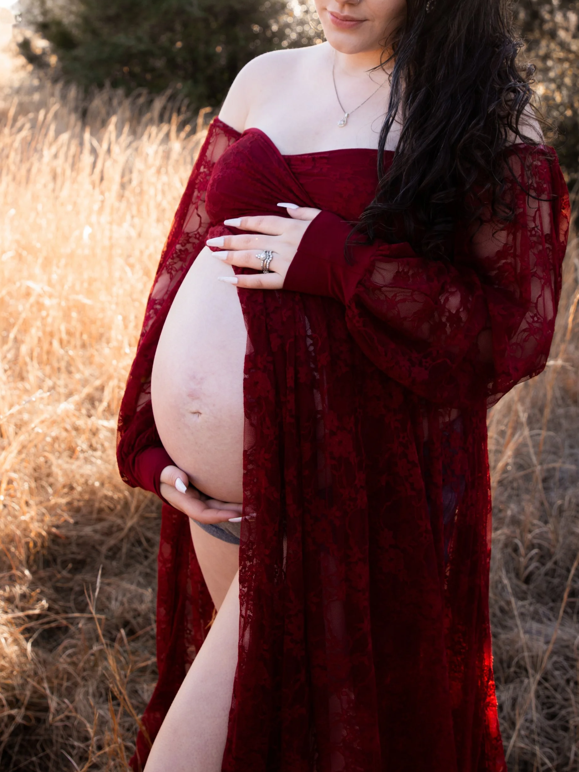 A woman named kaitlyn who is the best doula near me for birth at a hospital, birth center, or home with long dark hair wearing a red lace off-the-shoulder dress, holding her pregnant belly outdoors in a field with dry grass, during the golden hour.