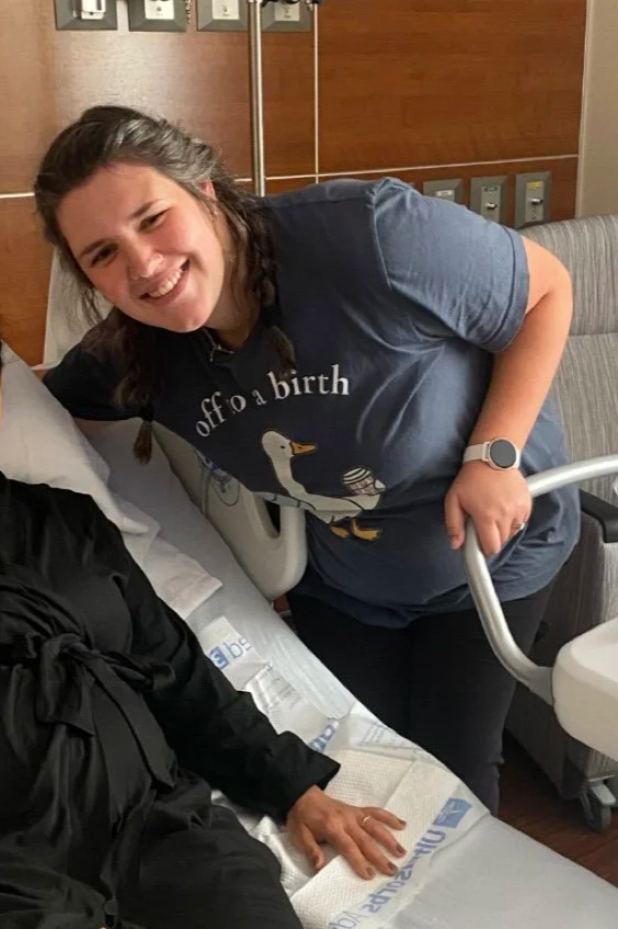 Smiling birth doula in Bryan Texas leaning over hospital bed with a client, holding onto a baby's hospital bed rail, in a hospital room after a birth.