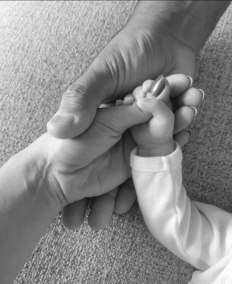 A close-up of an adult hand holding a baby's tiny hand after being supported by Kaitlyn Rosenbarker the best doula near me. The baby is wearing a long-sleeve shirt, and the adult's hand is gently grasping the baby's fingers.