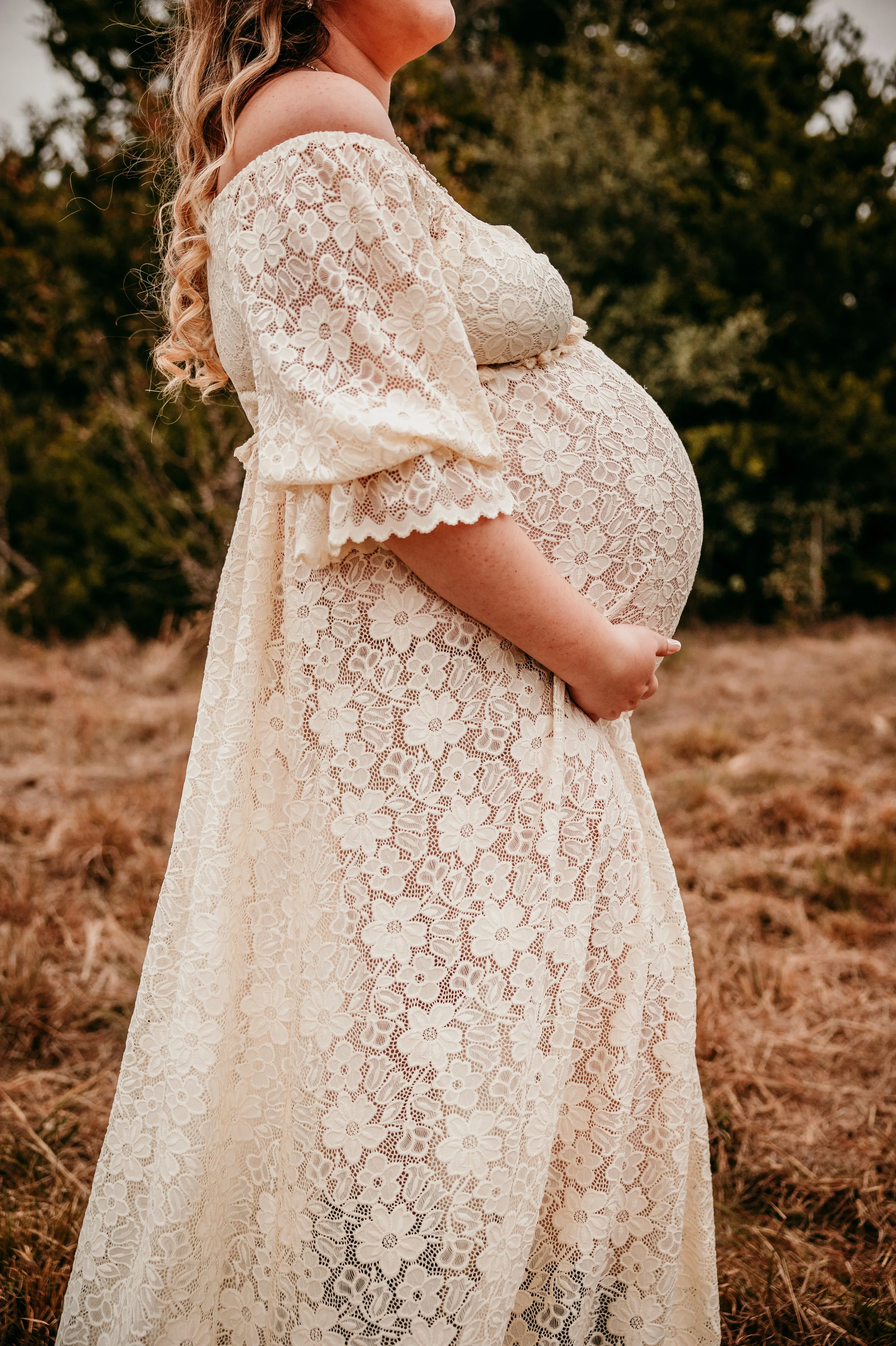 Pregnant woman named kaitlyn rosenbarker that owns light of life birth and is a good doula near me in bryan texas with availability for pregnancy and birth support wearing a white lace dress outdoors, with trees in the background.