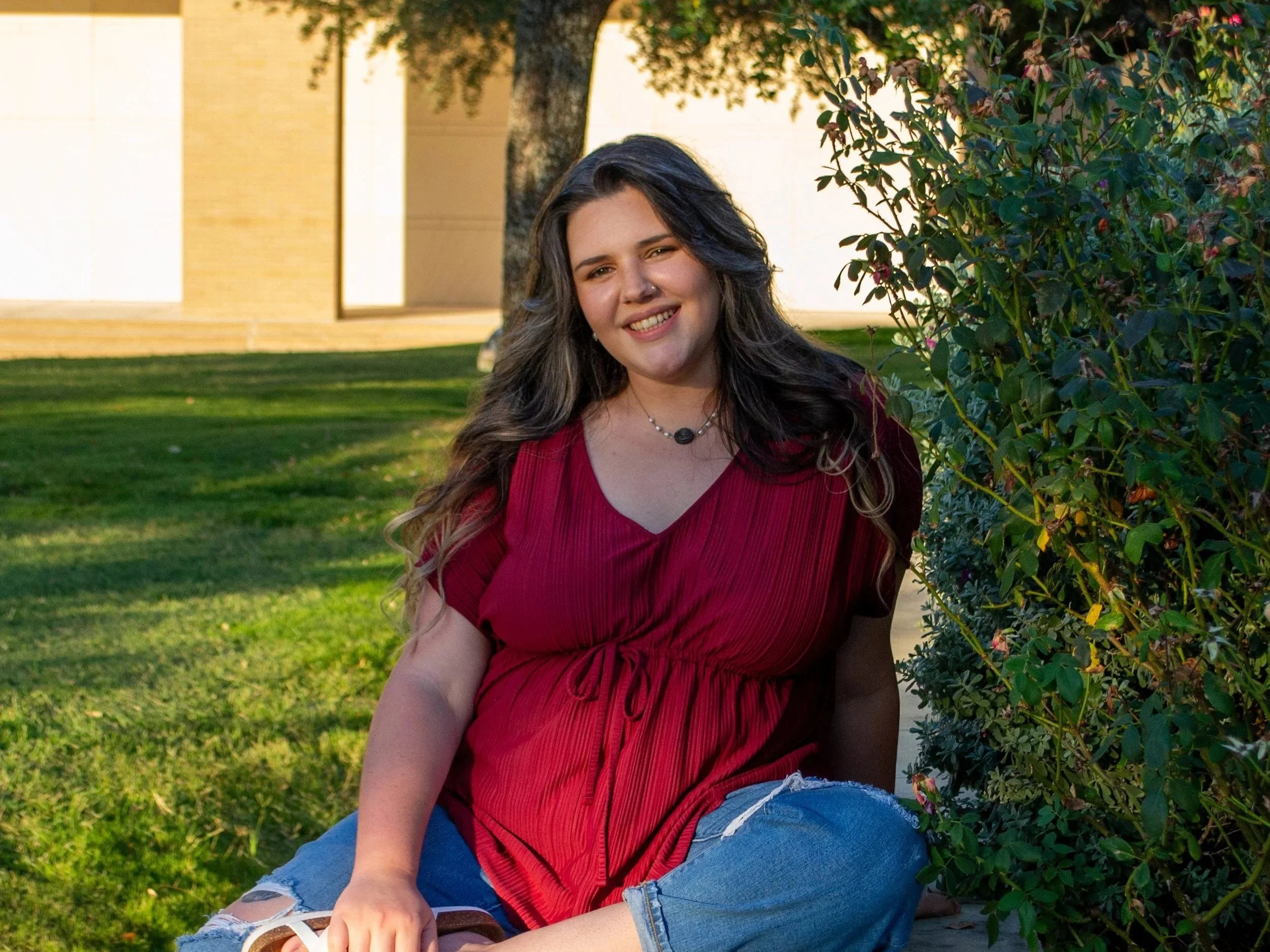 The Bryan College Station doula Kaitlyn Rosenbarker with long, wavy dark hair sitting on grass next to a bush with pink flowers, smiling at the camera, wearing a red top and ripped jeans, with a background of a tree and a building in the sunlight.