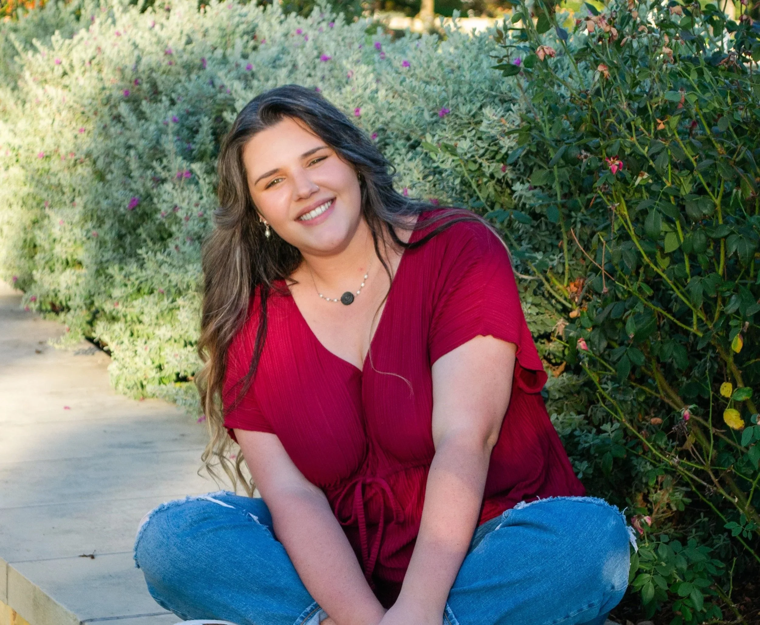 A doula in Bryan College Station Kaitlyn Rosenbarker with long dark hair and earrings, smiling, wearing a red top and ripped jeans, sitting outdoors near green bushes with pink flowers.