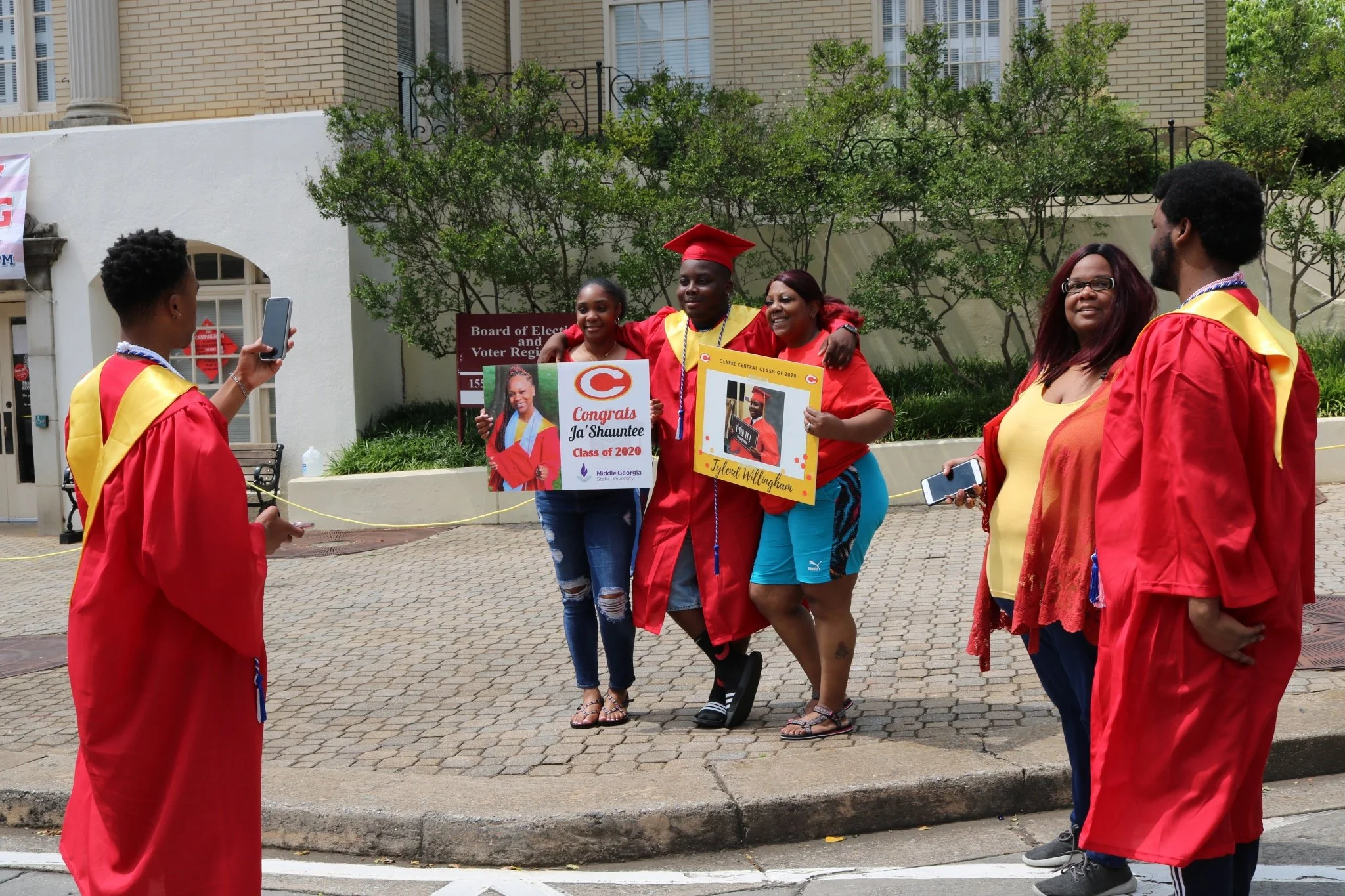 Group of five people celebrating graduation outdoors, with three holding signs and one taking a photo, all wearing red graduation gowns.