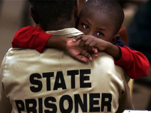 A young boy hugs a person wearing a white jacket with "STATE PRISONER" printed on the back, looking directly at the camera.