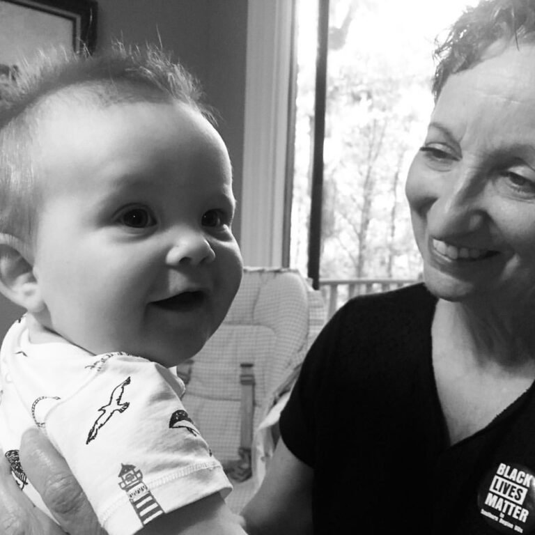 A smiling baby and woman interacting indoors near a window, with trees visible outside.