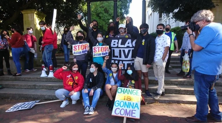 A group of diverse people protesting outdoors, wearing masks, holding signs that read "STOP Asian Hate" and "CHANGE IS GONNA COME," with a large "Black Lives Matter" banner, some raising fists in solidarity.