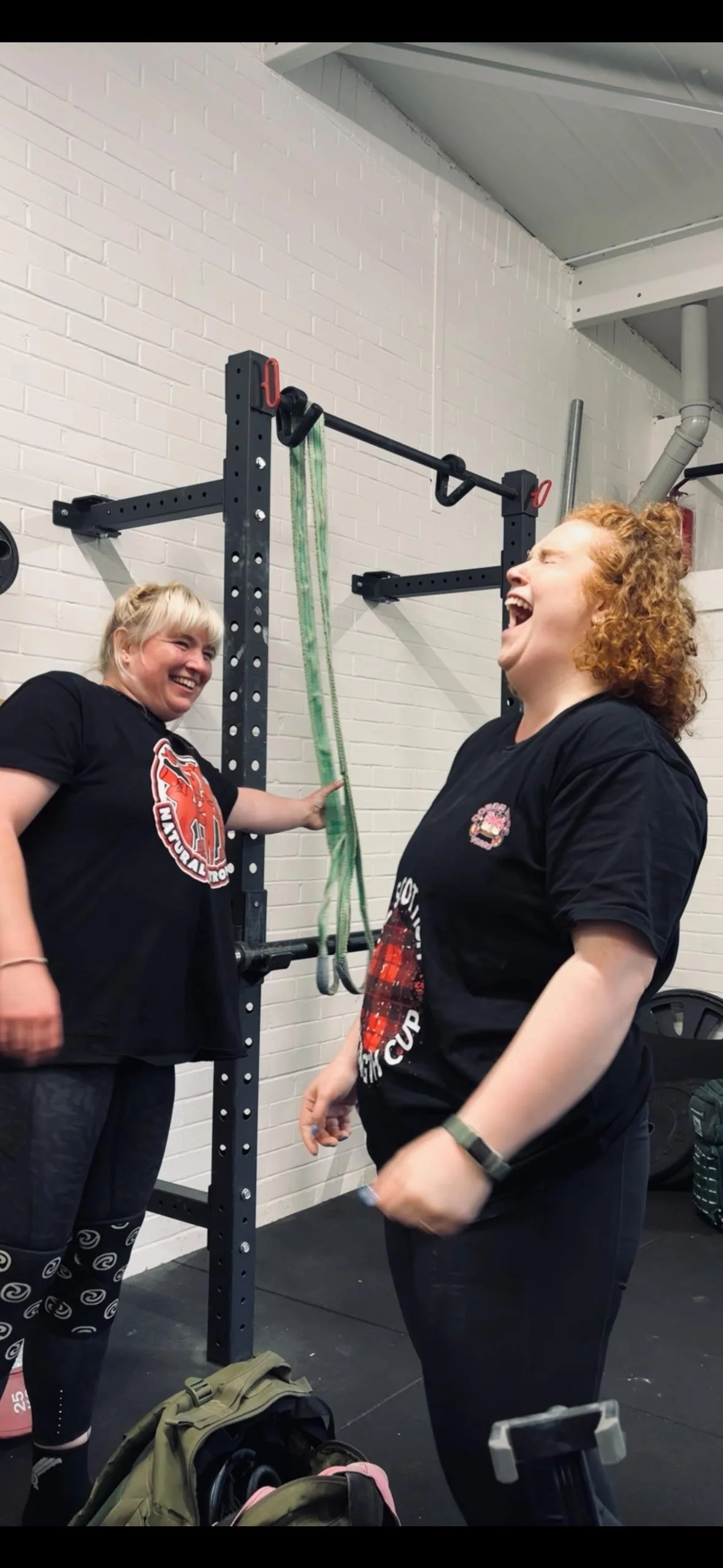 Two women laughing and smiling at each other in a gym, standing next to a squat rack with resistance bands hanging from it.