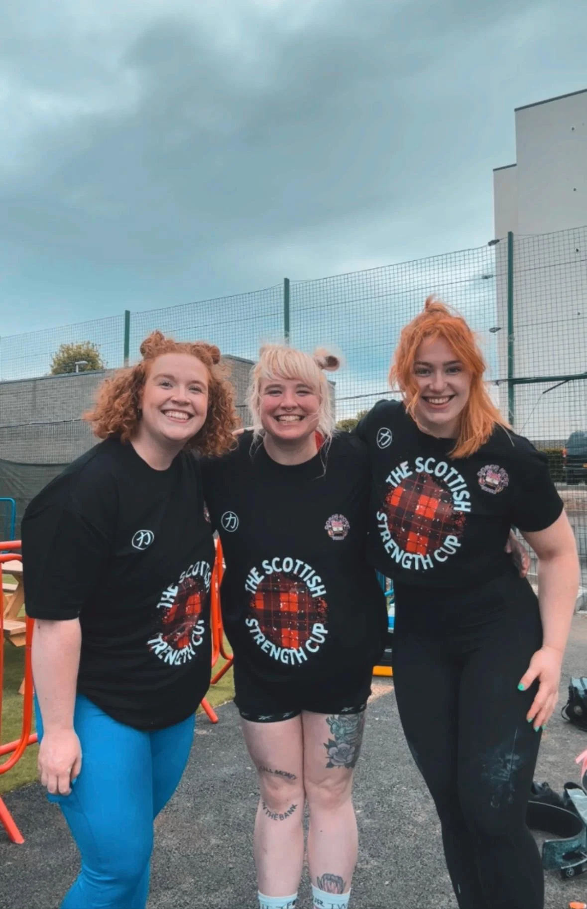 Three women smiling and posing outdoors, wearing matching black T-shirts with a Scottish tartan circle and the words 'The Scottish Strength Cup', standing in front of a fence on a cloudy day.