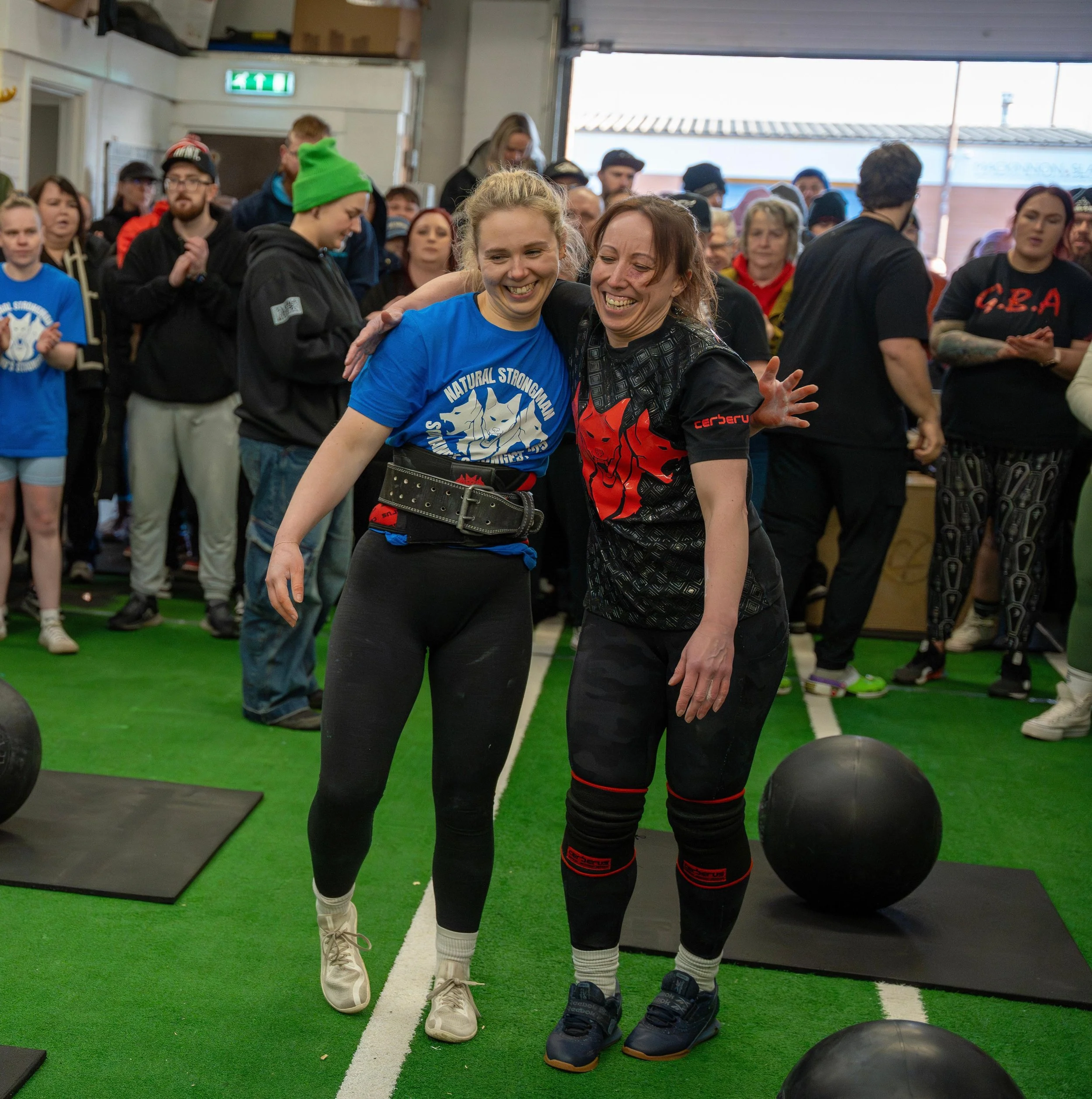 Two women sharing a happy moment at a strongwoman event, with one woman wearing a blue shirt and the other in black with red accents, surrounded by a crowd indoors.