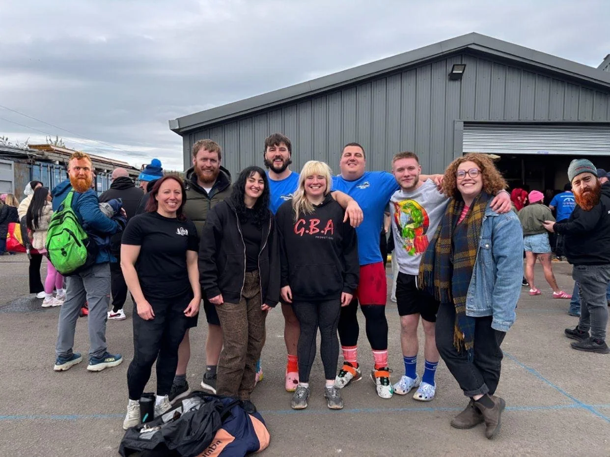 Group of nine people smiling and posing together outdoors, some in athletic wear, near a gray building and other people in the background
