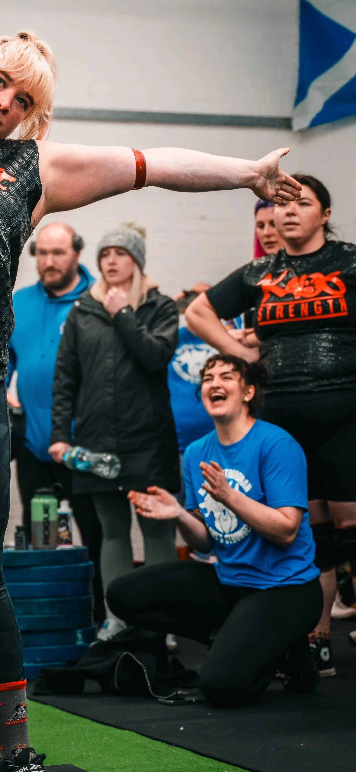 A woman in a blue shirt kneeling and smiling, surrounded by people in an indoor gym, with one woman pointing and others watching and reacting.