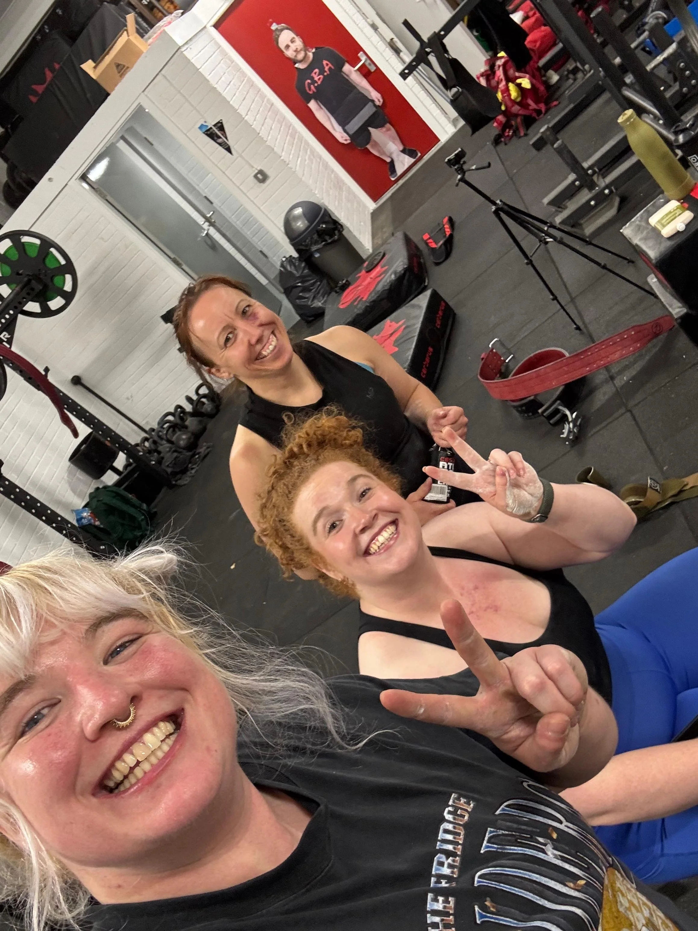 Three women smiling and posing for a selfie in a gym. The woman in the front has platinum blonde hair, a septum piercing, and is wearing a black t-shirt. The woman in the middle has curly red hair and is making a peace sign. The woman in the back has