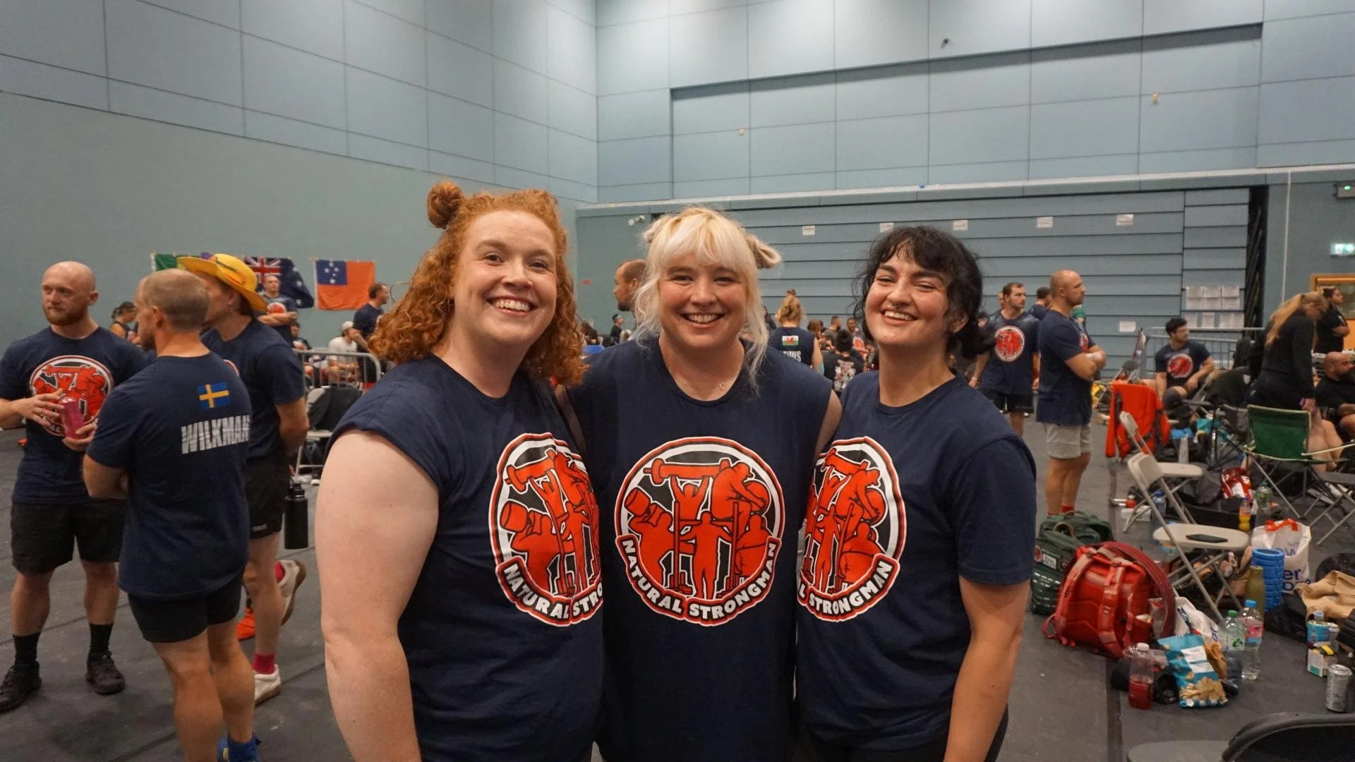 Three women wearing matching navy blue T-shirts with a logo, smiling and posing during an indoor indoor sports event, with other participants and flags in the background.