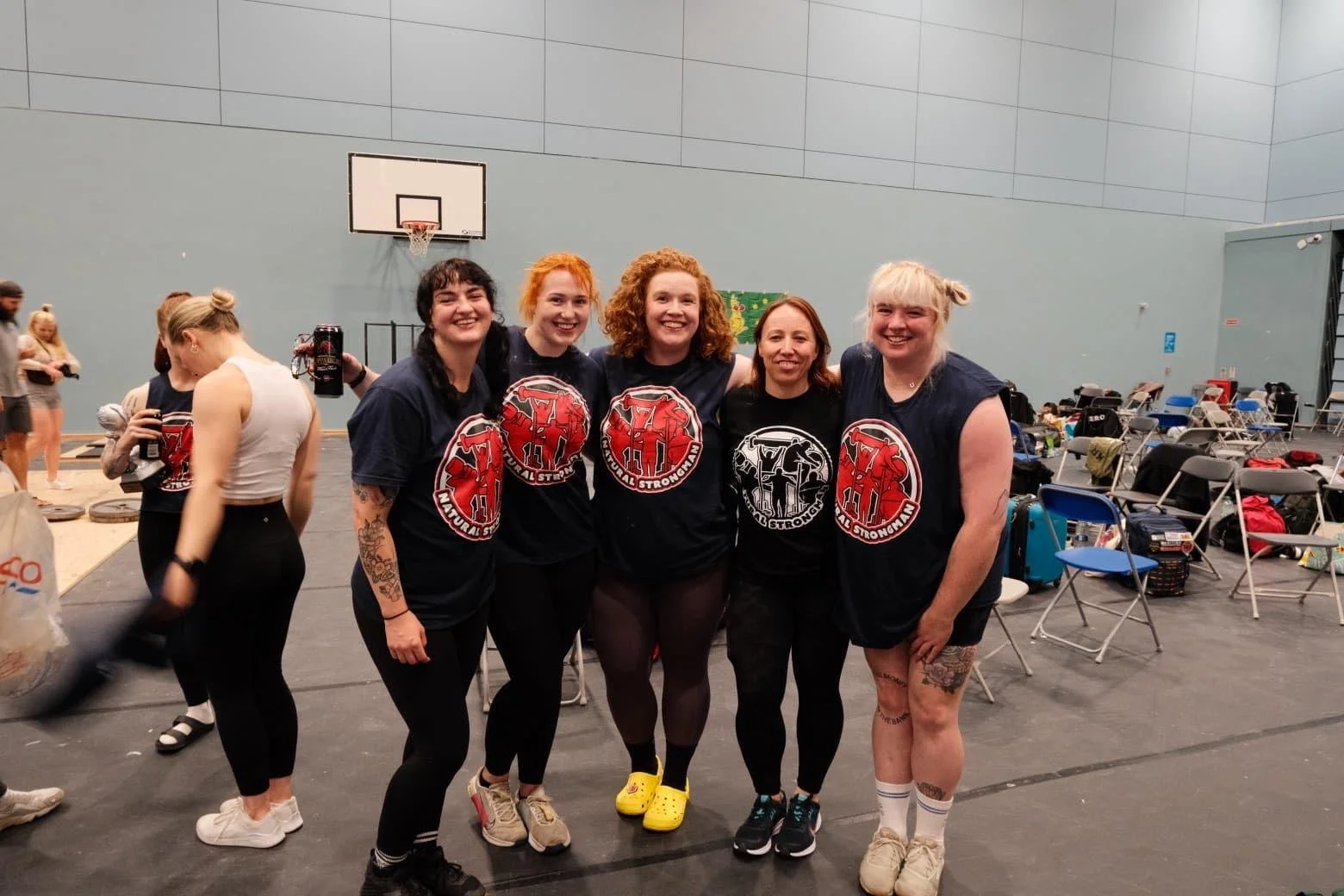 Group of five women in athletic clothing smiling indoors, surrounded by chairs and gym equipment.