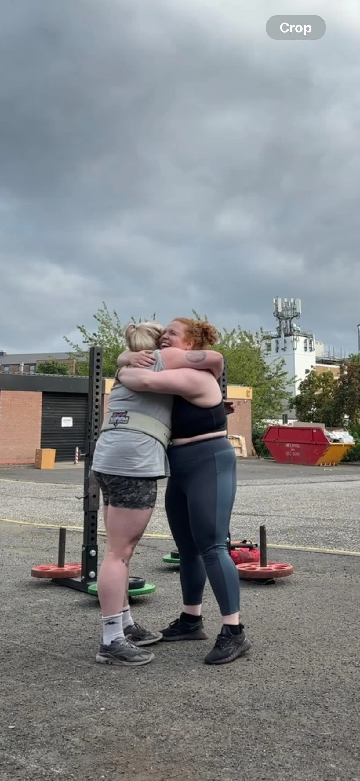 Two women hugging outdoors on a cloudy day, standing on gravel with weightlifting equipment behind them