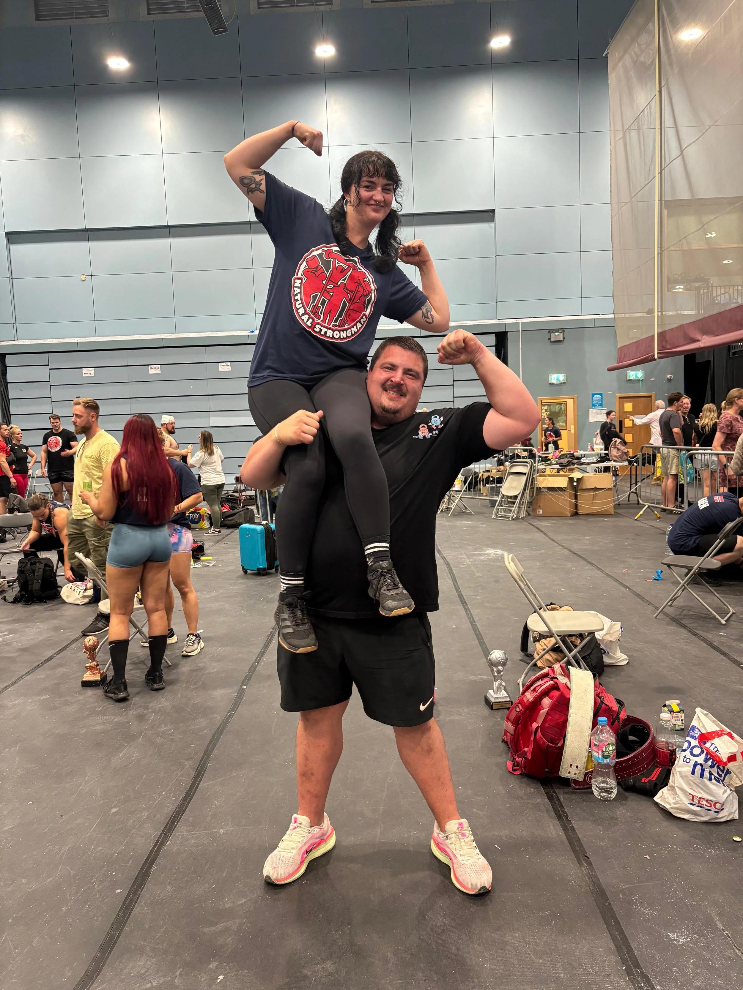 A woman sitting on a man's shoulders, flexing her arms in a gym setting. The man is standing with his arms also flexed. Both are smiling. The background shows other people, some with trophies, and gym equipment.
