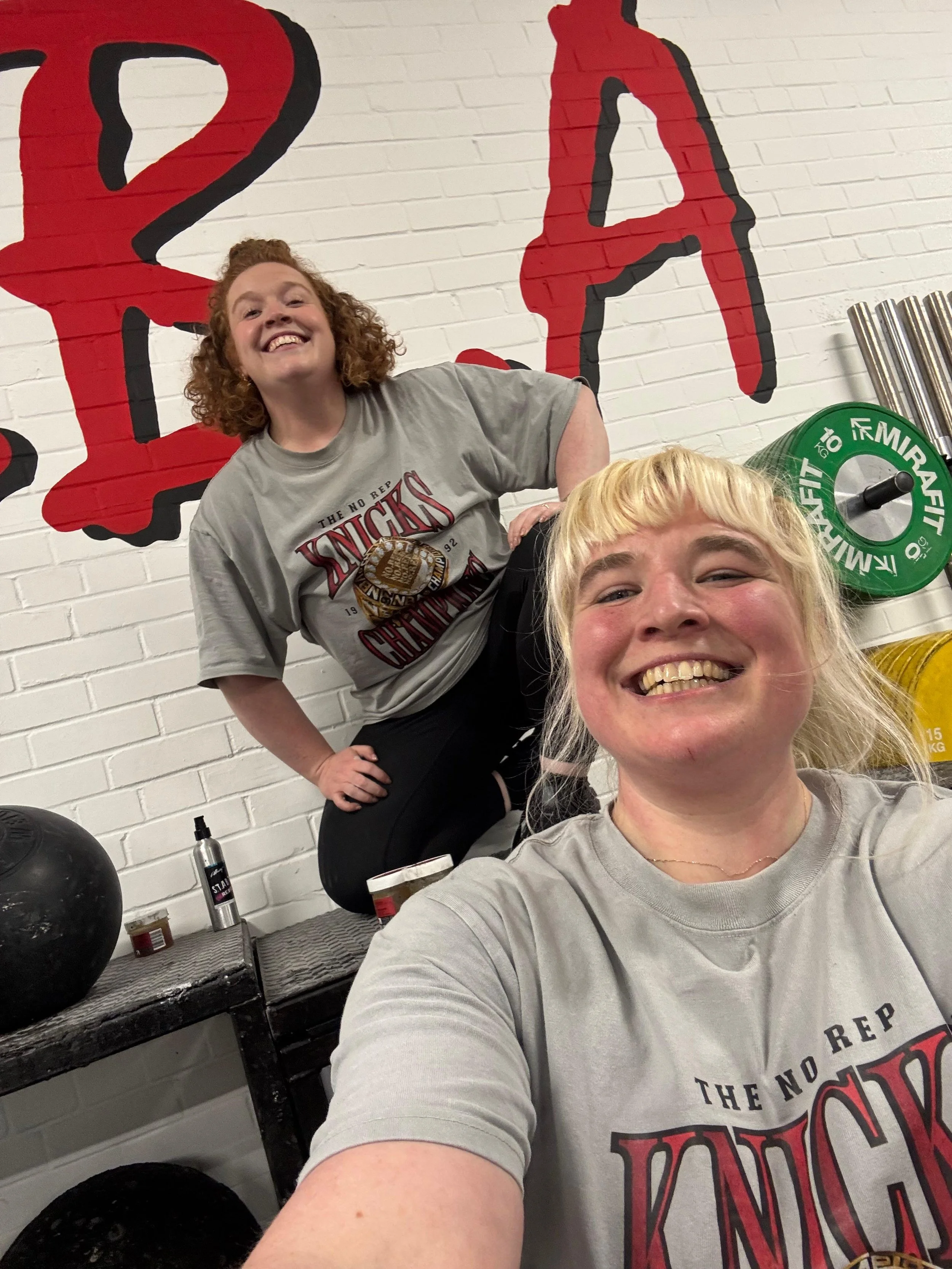 Two smiling women in a gym taking a selfie. One woman with curly red hair standing behind another woman with blonde hair. Both are wearing gray t-shirts with a sports logo.