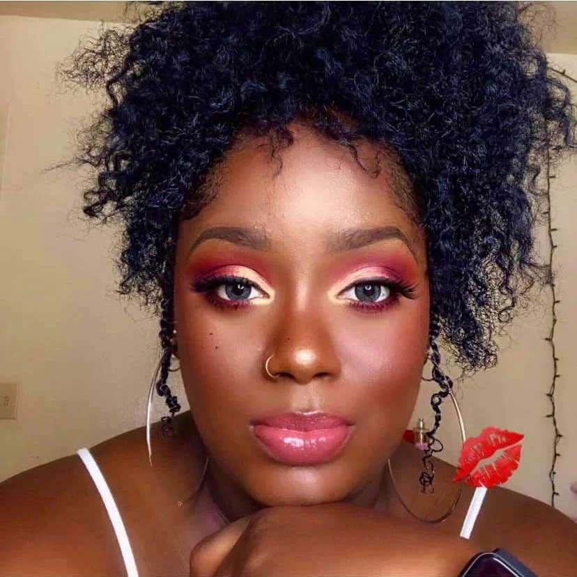 Close-up portrait of a woman with curly black hair, bright makeup including pink eyeshadow, eyeliner, and glossy lips, wearing hoop earrings with red beads, a nose ring, and pink lipstick, resting her chin on her hand.