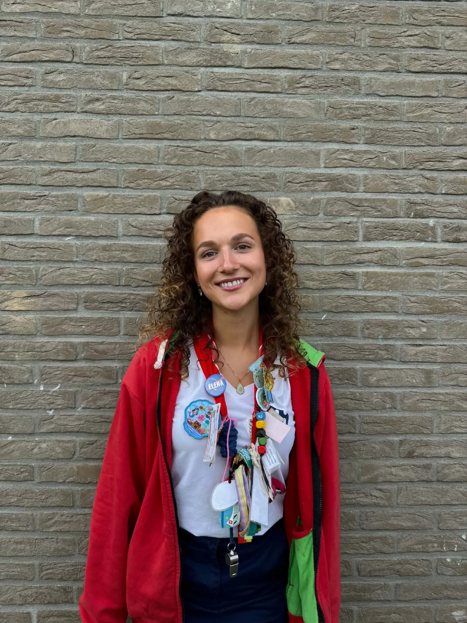 A woman smiling in front of a brick wall, wearing a white shirt with a logo, a red jacket with green accents, and lots of colorful medals and ribbons around her neck.