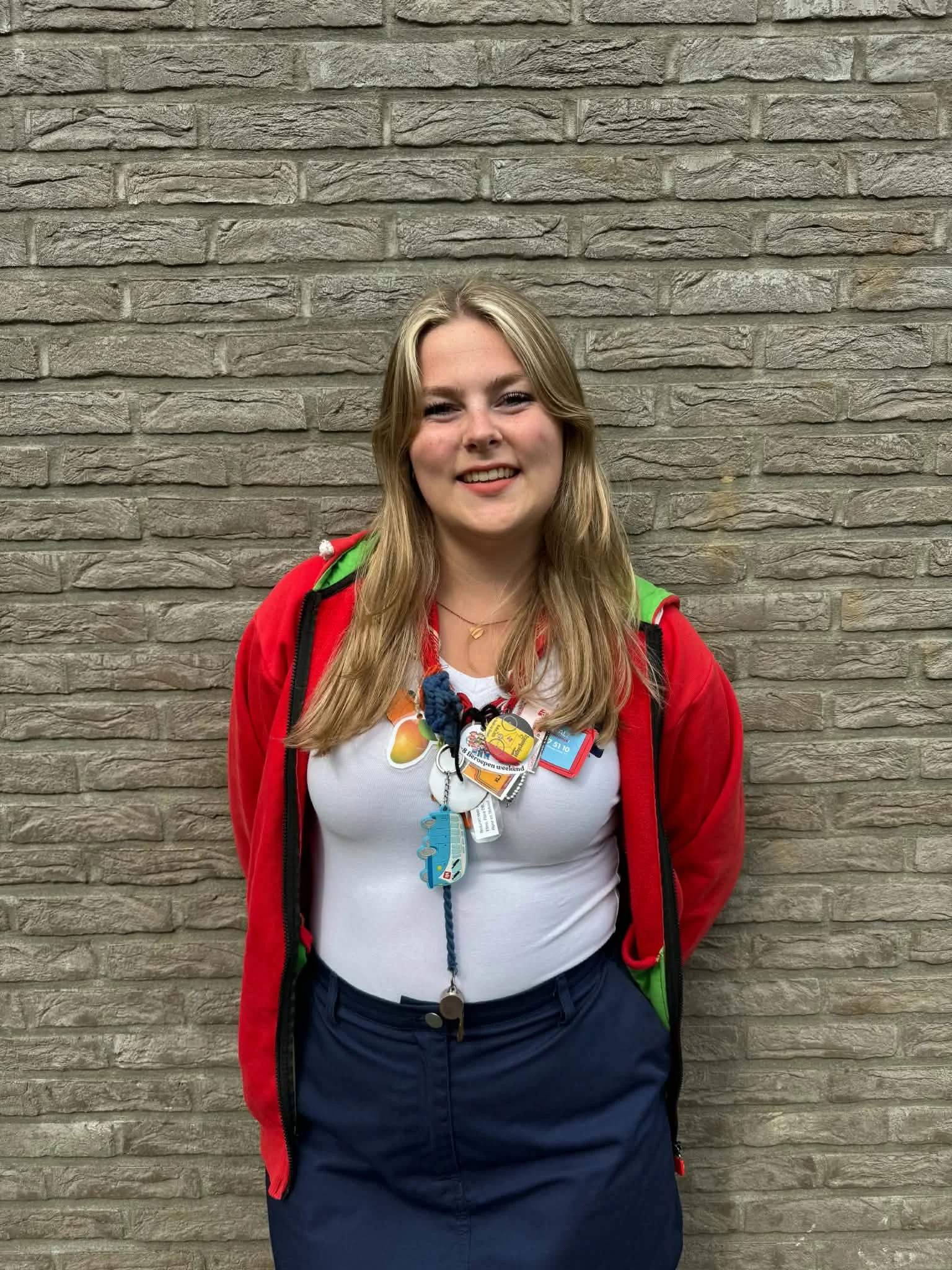 A young woman with long, wavy blonde hair posing in front of a brick wall. She is wearing a red jacket over a white shirt and has a whistle hanging from a blue braided lanyard around her neck.