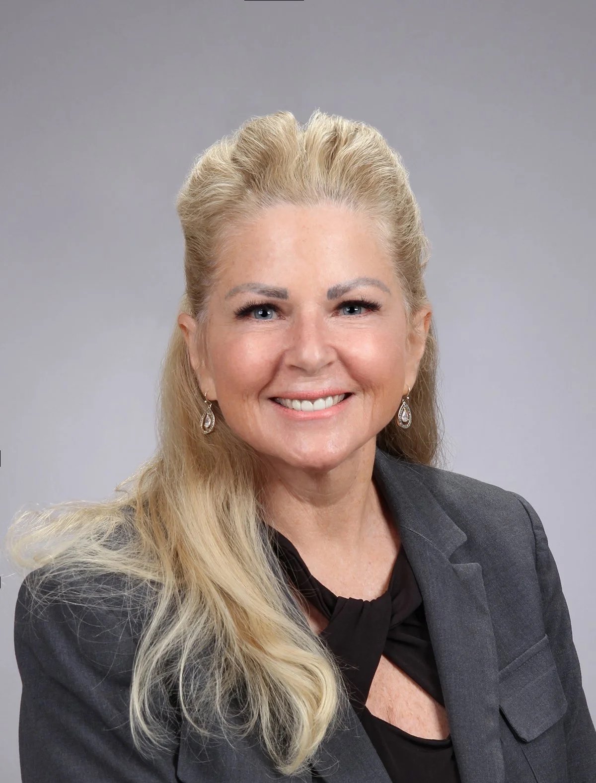 Portrait of a smiling woman with blonde hair, blue eyes, wearing a dark blazer and earrings, against a gray background.
