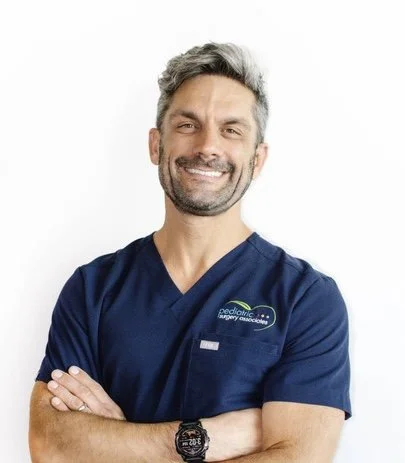 Smiling young woman with shoulder-length black hair, wearing a blue top, standing among green foliage in a greenhouse or botanical setting.