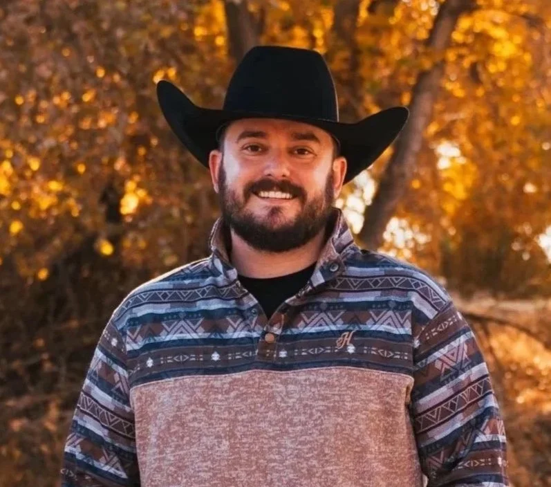 A man smiling outdoors wearing a black cowboy hat and a patterned southwestern style shirt with brown, black, and blue colors, with fall foliage in the background.