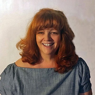 A man with curly dark hair, glasses, and a beard, sitting indoors near a plant.