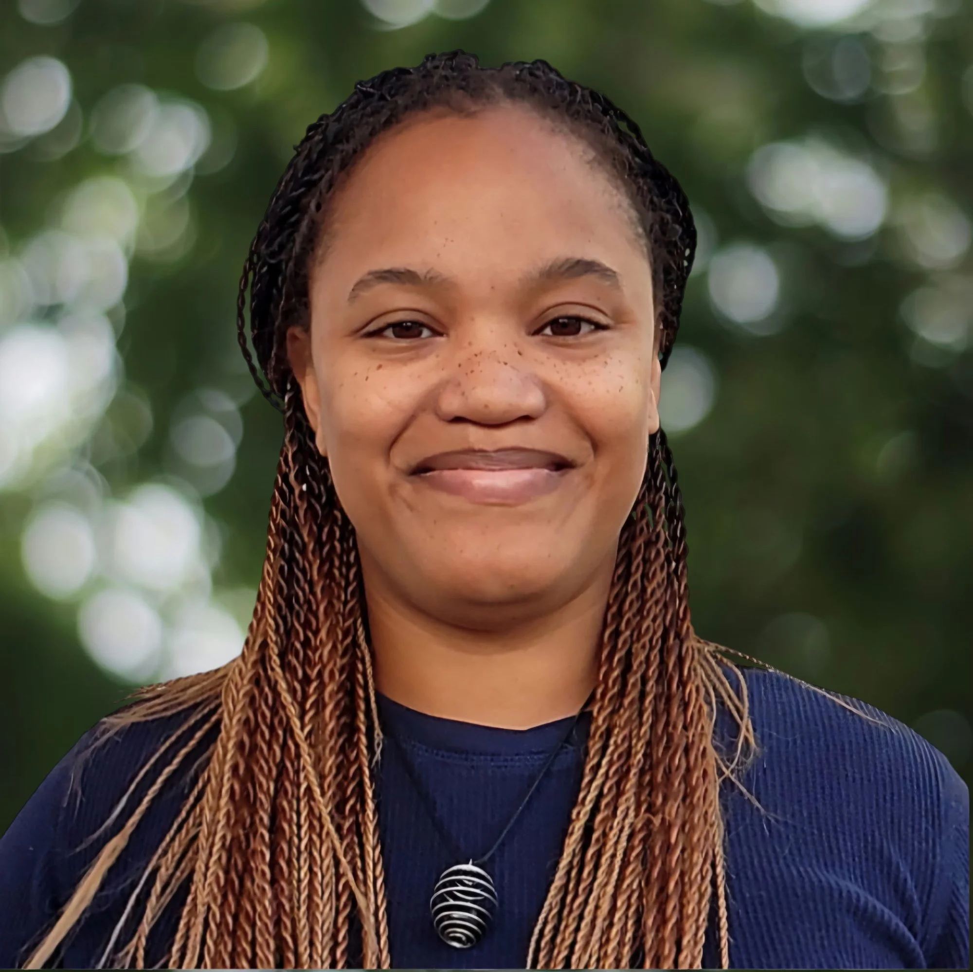 Close-up of a woman with braided hair smiling outdoors with a blurred green background.
