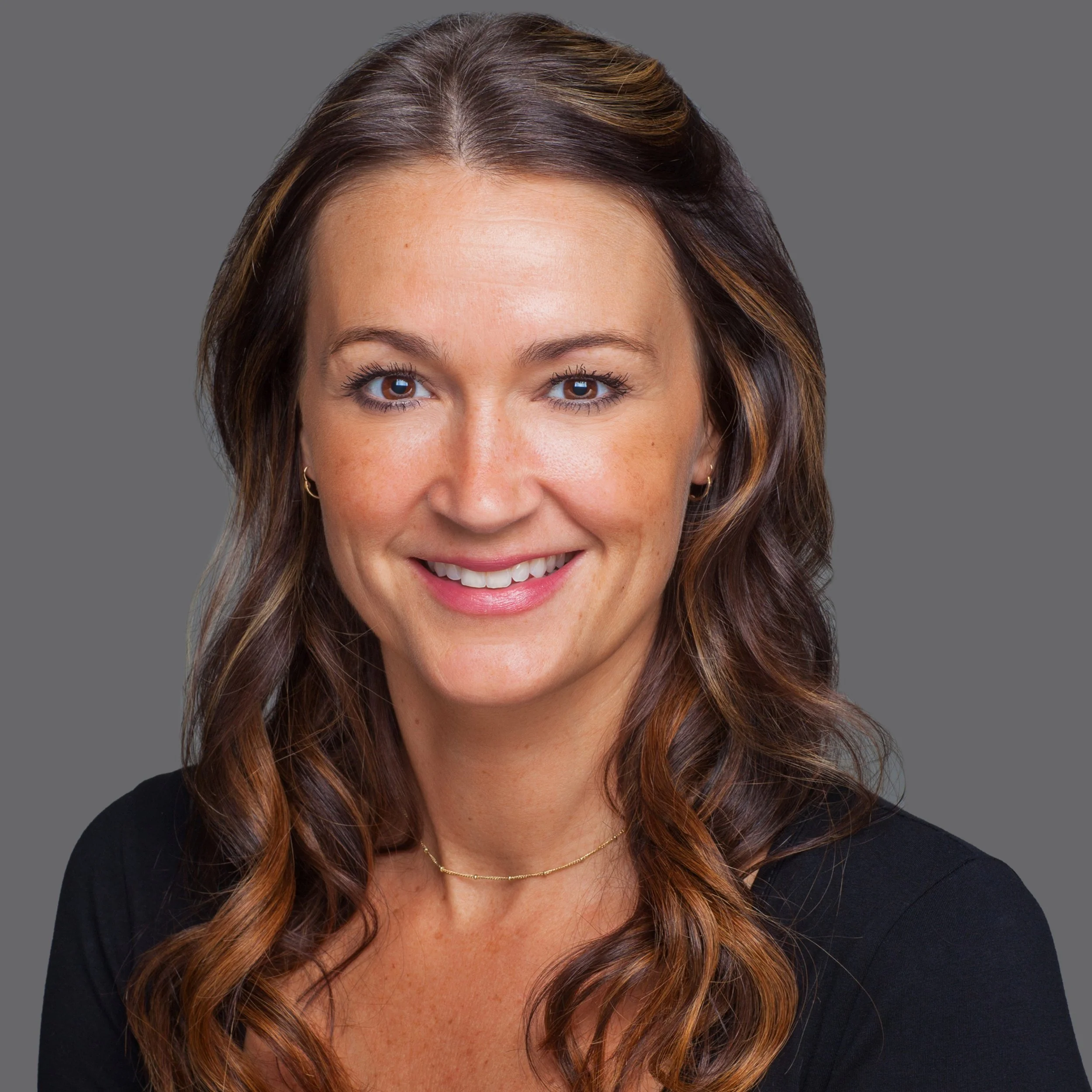 Headshot of a smiling woman with wavy brown hair, wearing a black top and gold necklace, against a gray background.