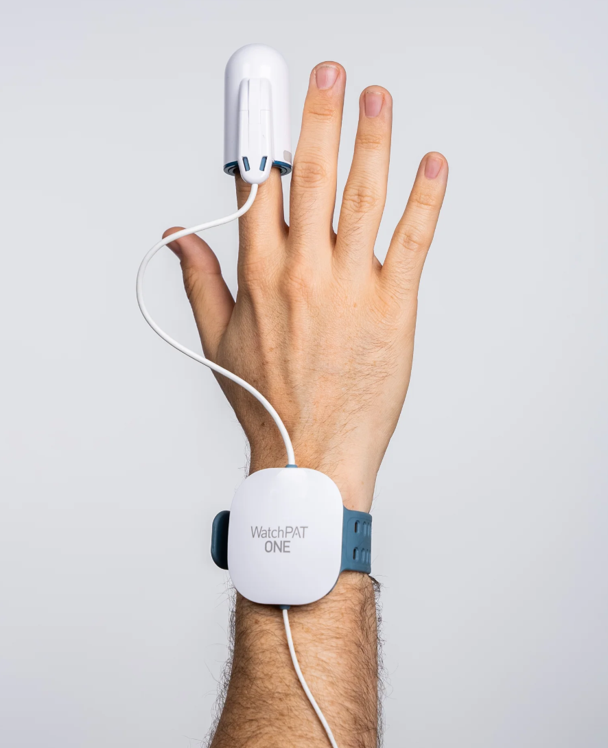 A person's hand with a white wrist-worn electronic device labeled WatchPAT ONE and a finger attached to a monitoring clip connected to a cylindrical sensor on the finger, against a plain white background.