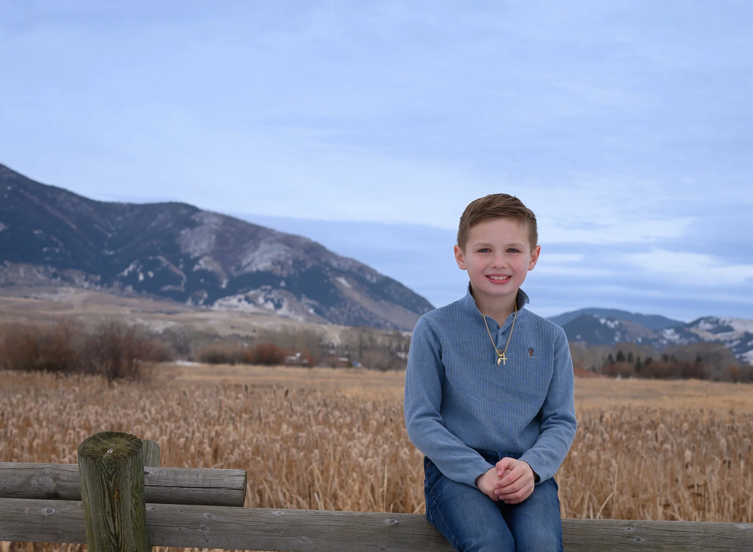 A young girl with red hair wearing a floral crown is outdoors in a grassy area with yellow flowers. Someone is touching her face with a pink flower.