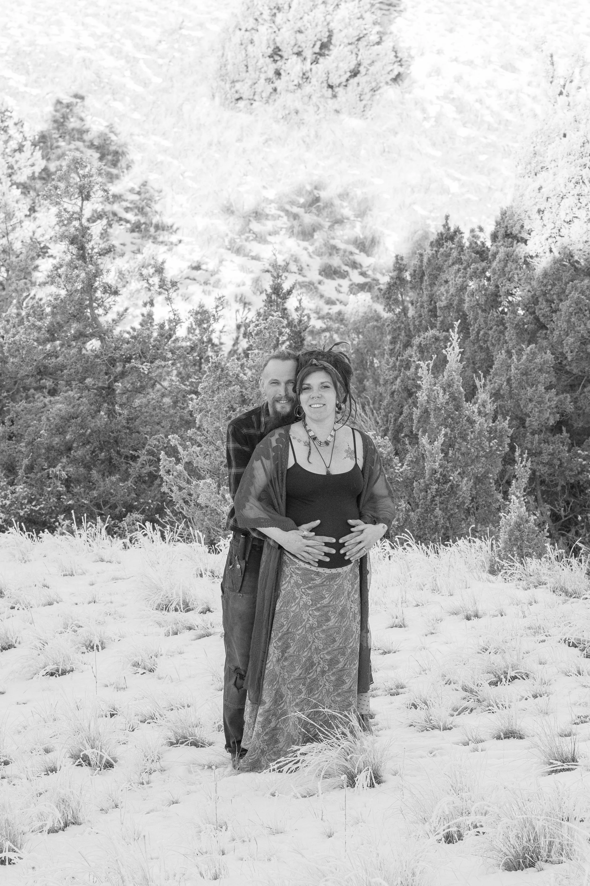 A black and white photo of a couple standing in a grassy field with trees and mountains in the background. The man is standing behind the woman, holding her belly. She is wearing a long skirt and a top with a jacket, and a necklace, while he wears a plaid shirt.