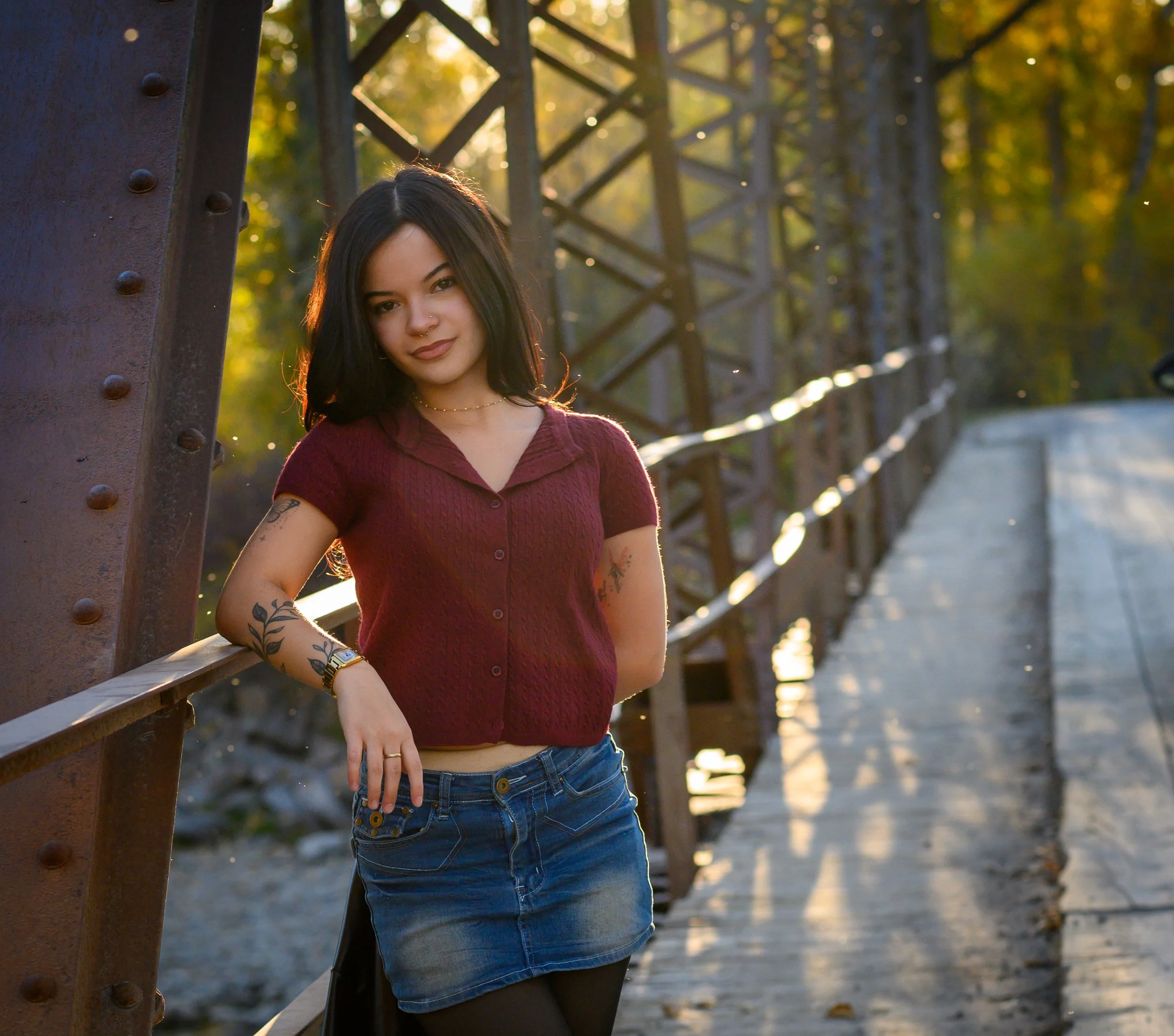 A young woman with black hair and tattoos, wearing a maroon short-sleeve button-up shirt, denim mini skirt, black tights, and black boots, standing on a bridge during fall with trees and a river in the background.