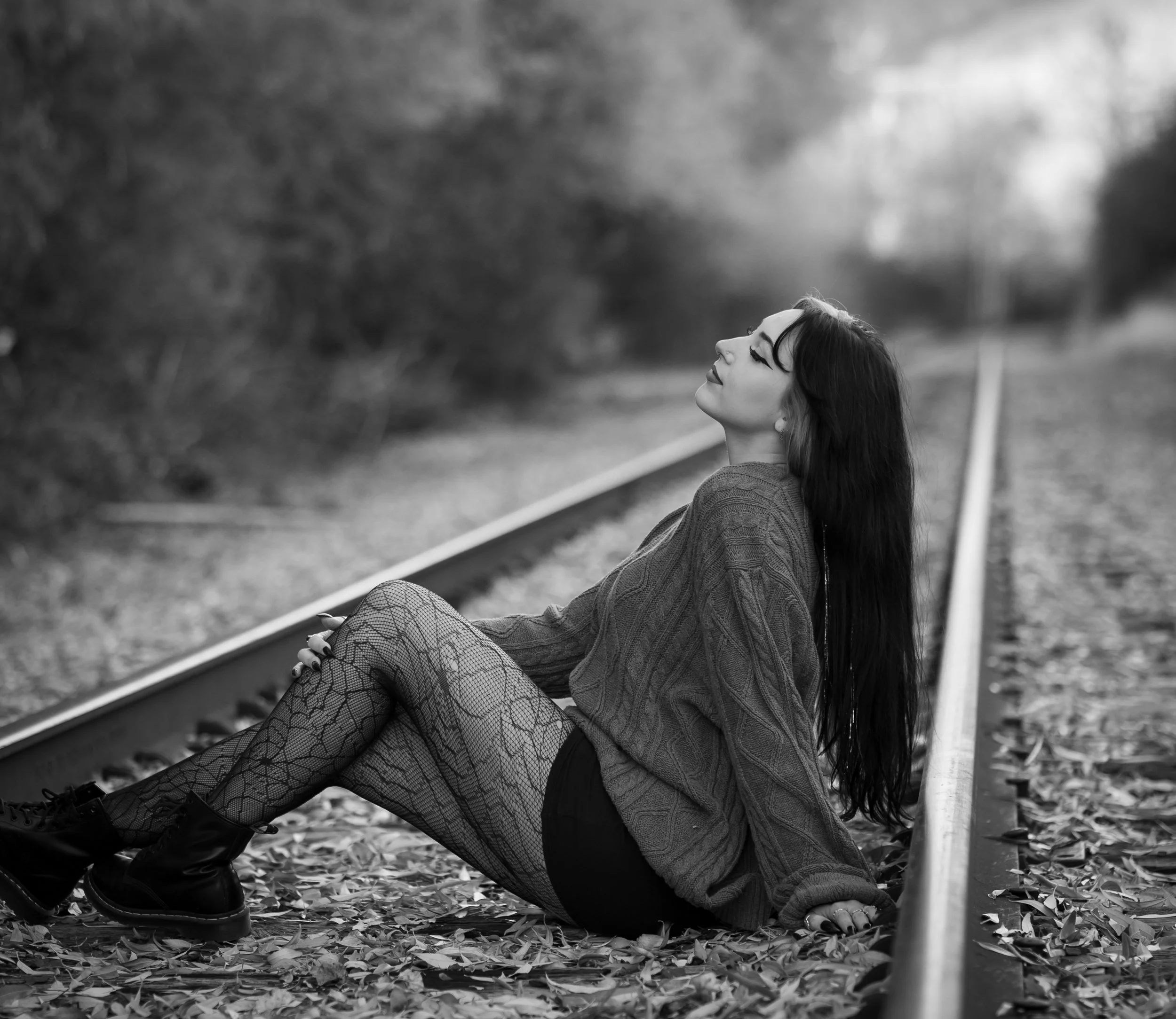 A young woman with black hair and tattoos, wearing a maroon short-sleeve button-up shirt, denim mini skirt, black tights, and black boots, standing on a bridge during fall with trees and a river in the background.