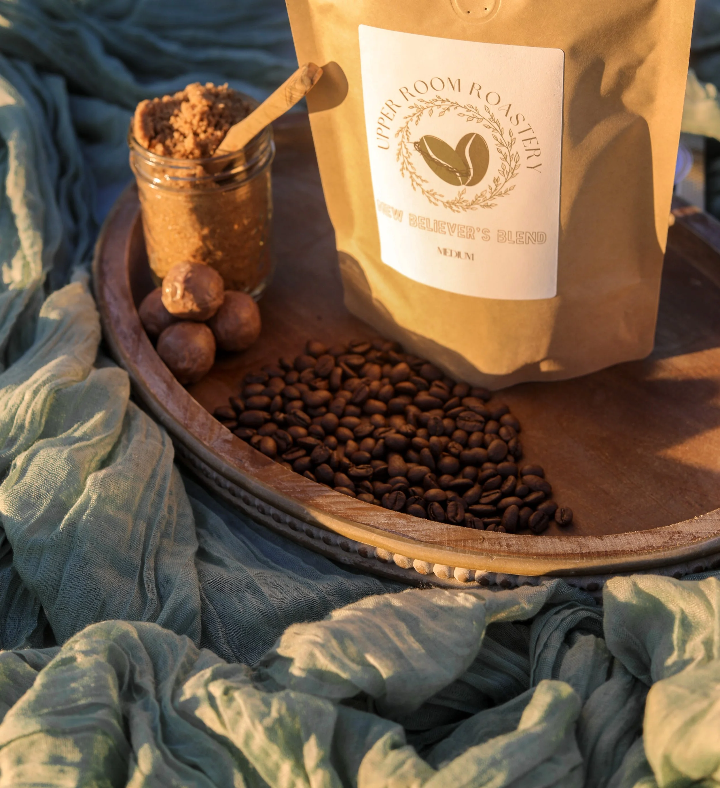 A wooden tray with a bag of Upper Room Roastery coffee, a jar of dark brown sugar, fresh medium roasted coffee beans, and some milk chocolate truffles, all surrounded by green fabric.
