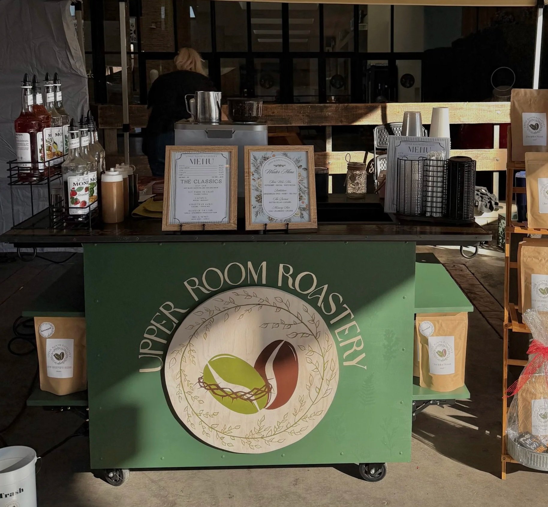 A mobile coffee and snack stand with a green front labeled 'Upper Room Roastery,' displaying coffee bags, condiments, and menu signs within a rustic setting.