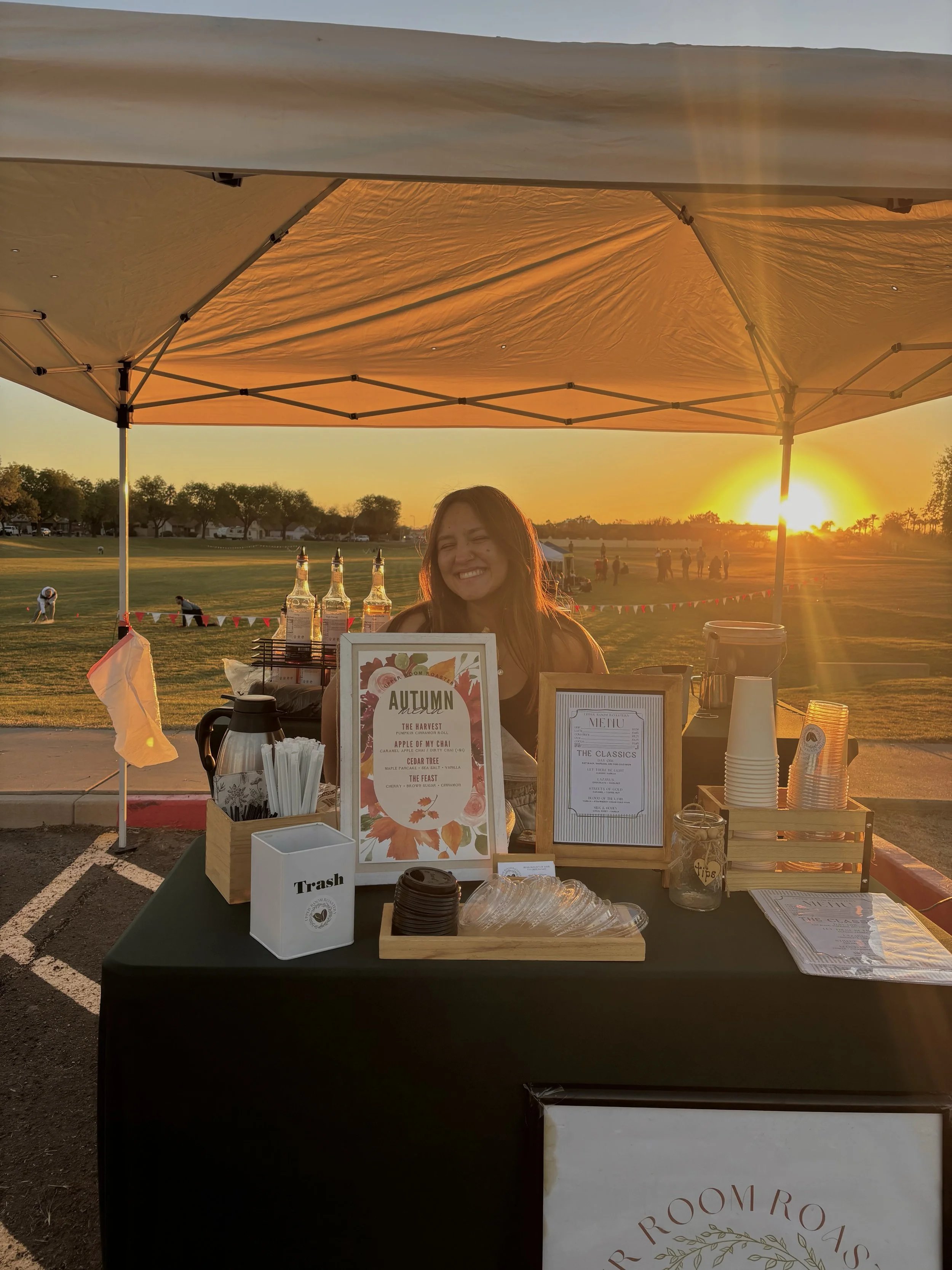 A woman smiling at a beverage stand during sunset, with bottles, cups, and signs on the table, set up outdoors in a park or open field.
