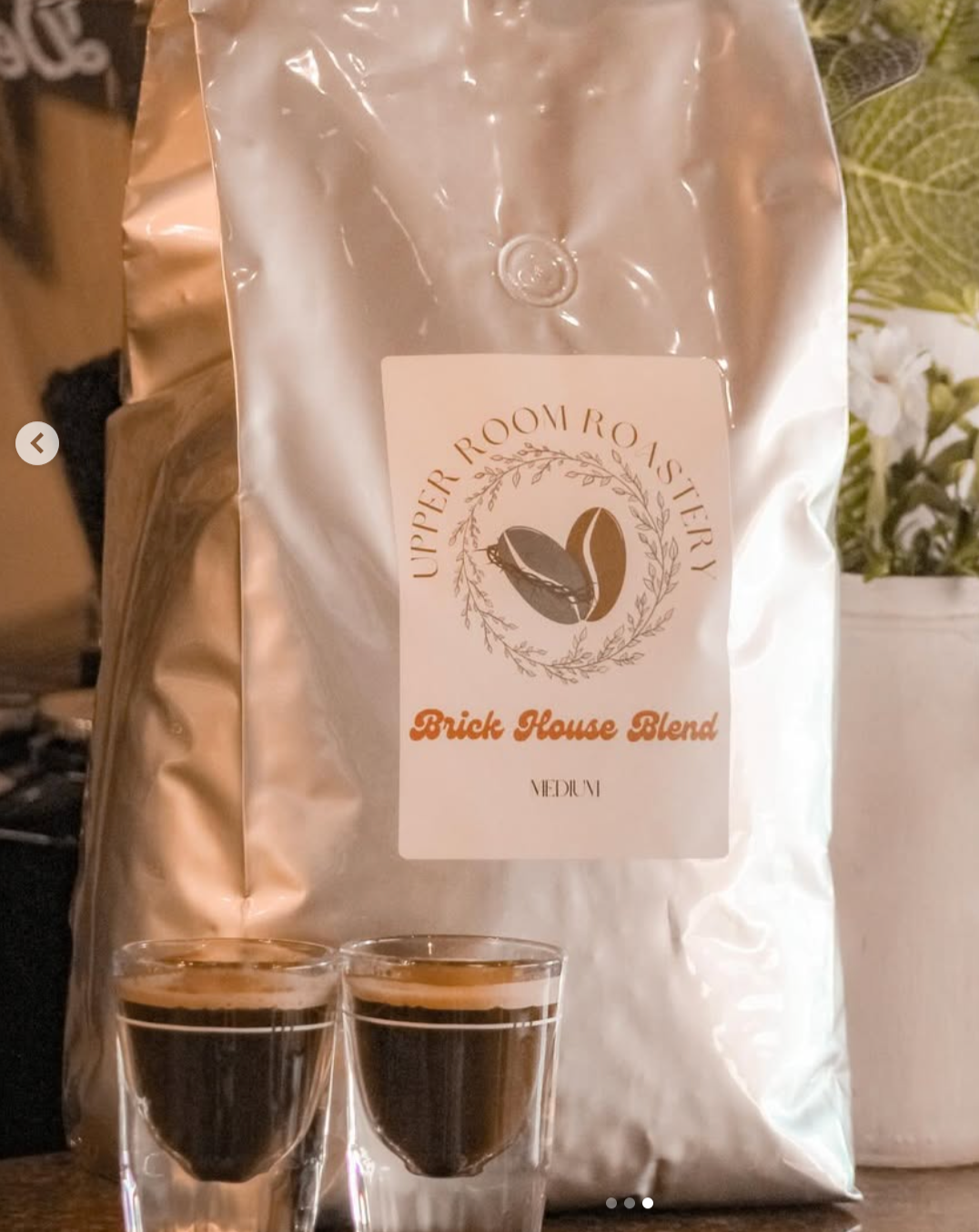 A silver bag of Upper Roov Roastery Brick House Blend coffee beans labeled medium roast, with two small glasses of black coffee in front of it.