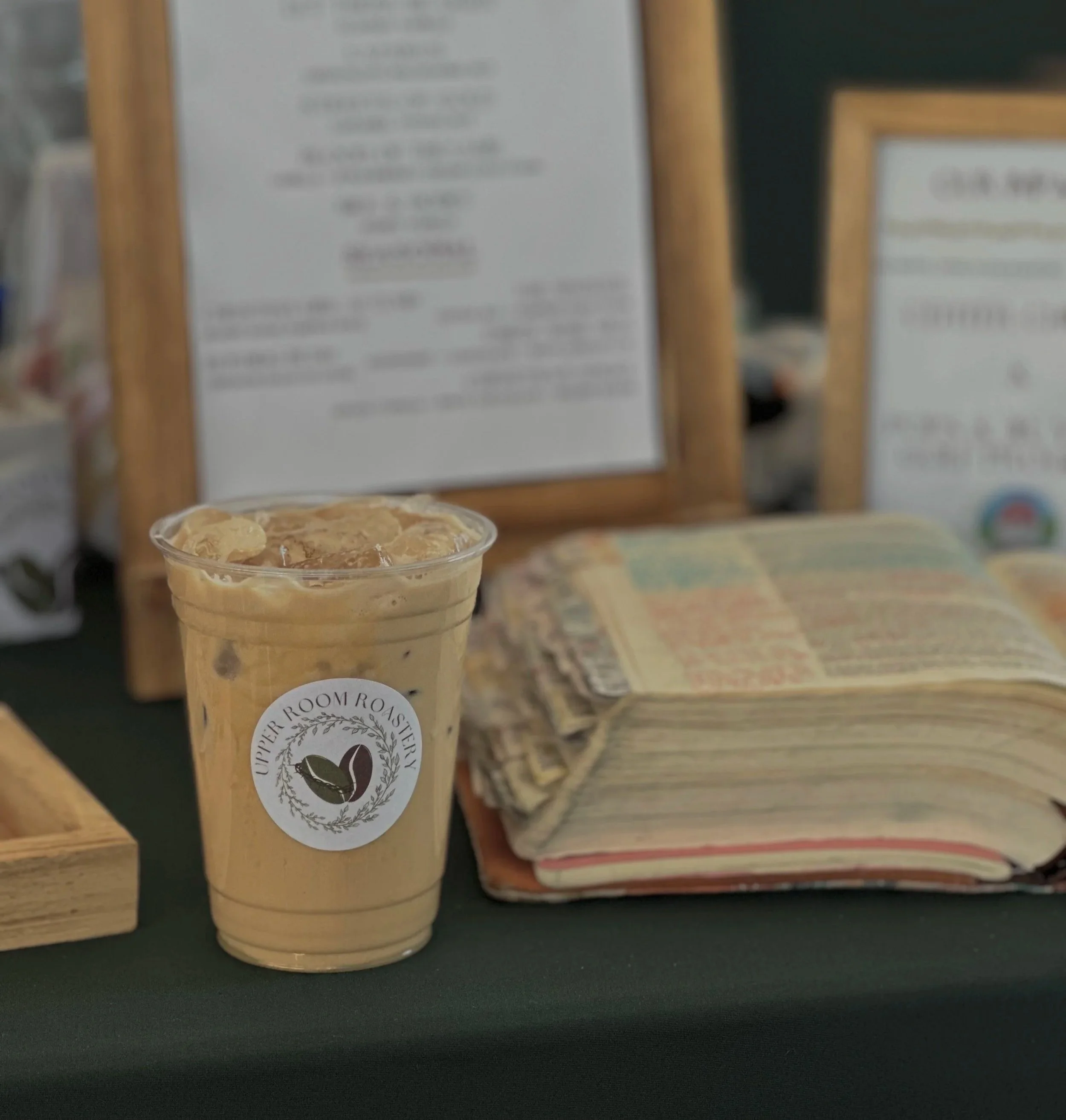 Iced coffee drink with ice cubes in a clear plastic cup with a logo for Upper Room Roastery, placed on a dark surface with a blurred background of framed posters and a stack of paper or receipts.