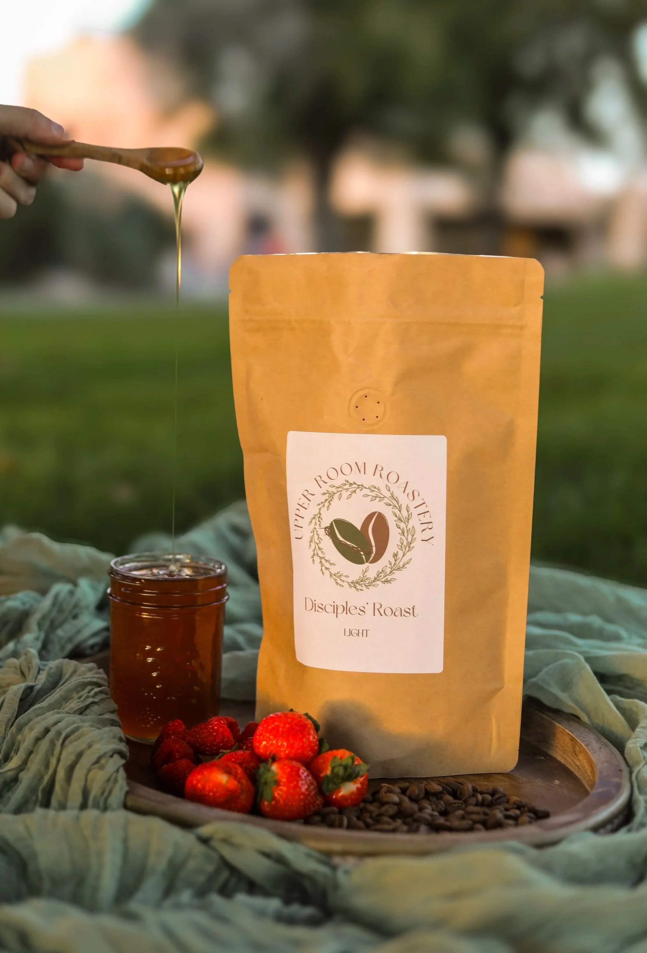 A package labeled 'Disciples’ Roast Light' from Upper Room Roastery with a spoon dripping honey above it, a small jar of honey, fresh strawberries, and roasted coffee beans on a wooden tray, set outdoors on a green cloth with trees in the background.