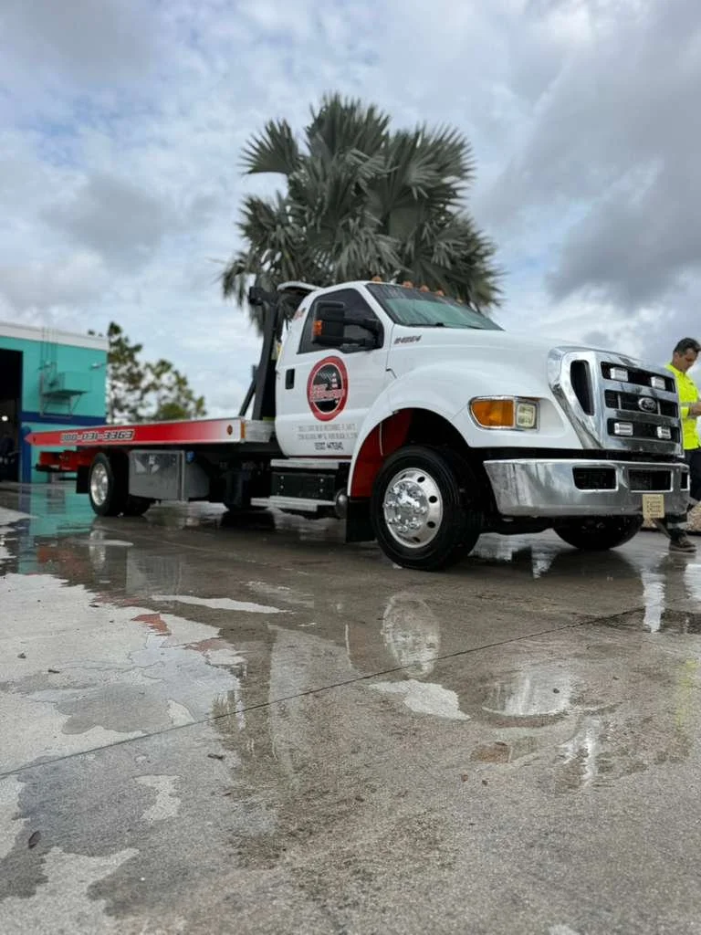 White tow truck parked on wet pavement with a palm tree, sky, and person in high-visibility jacket in the background.