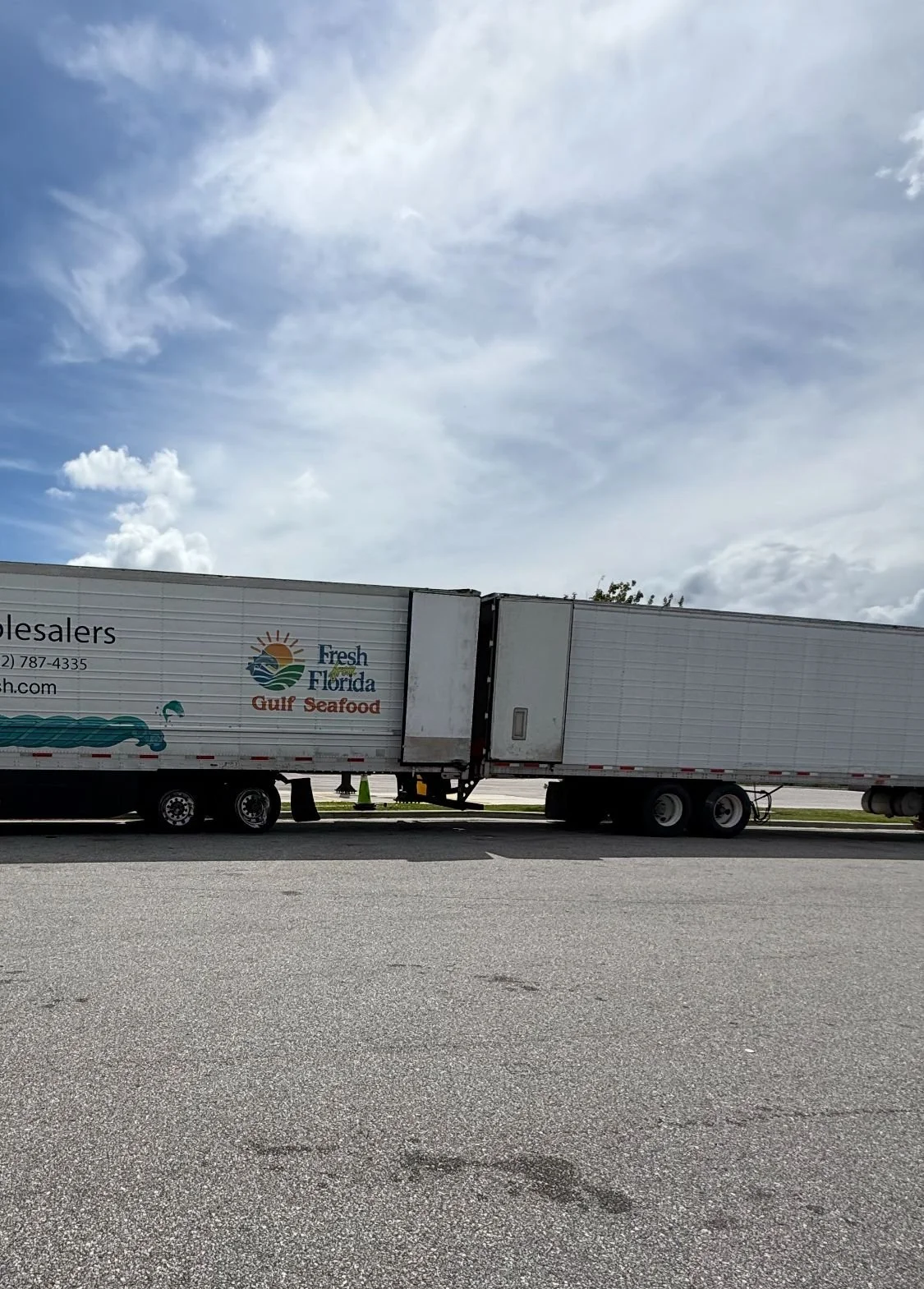 Large semi-truck with a white trailer parked on the side of the road under a blue sky with scattered clouds; the trailer has a logo and text for Fresh Florida Gulf Seafood.