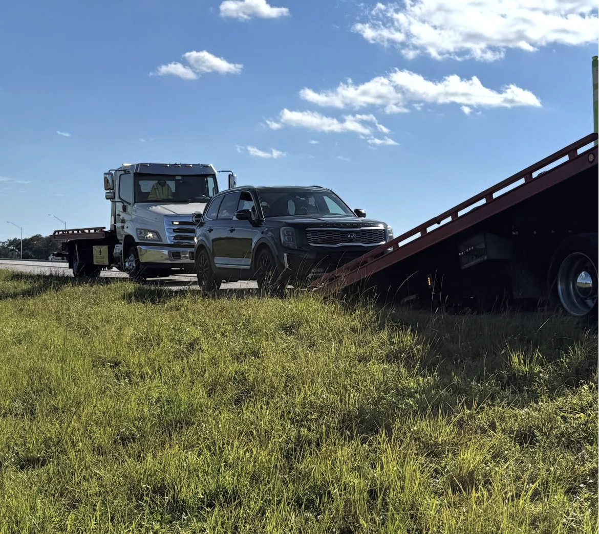 A tow truck is towing a black Kia SUV on the side of a highway under a blue sky with scattered clouds.