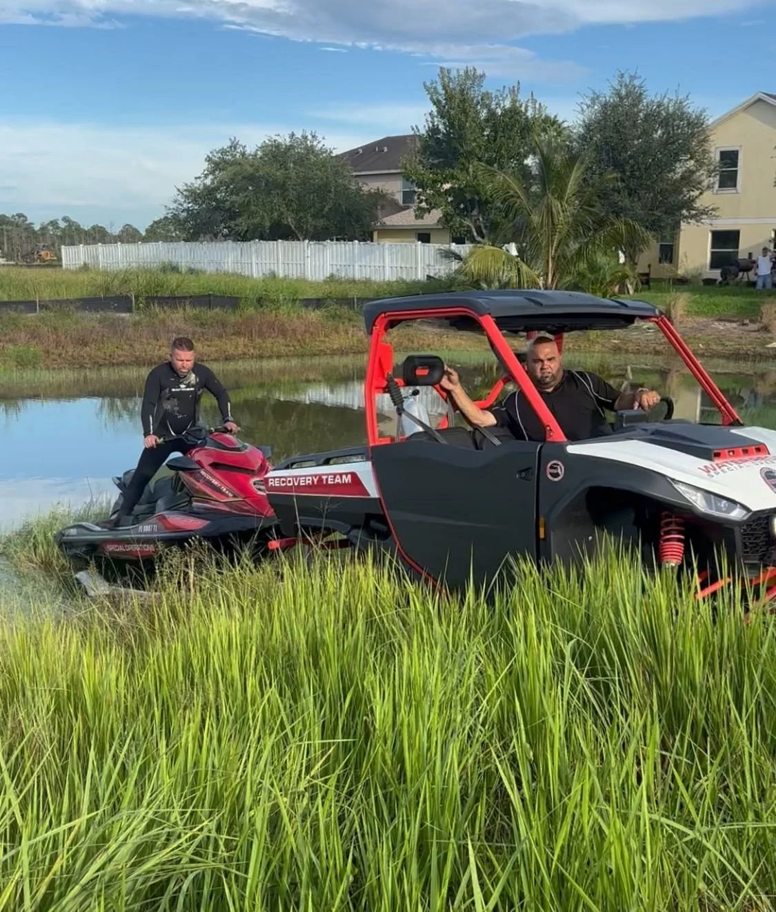 Two rescue team members equipped with emergency gear on a black and red rescue vehicle and a snowmobile near a small body of water, with tall green grass in the foreground and suburbs with houses and trees in the background.