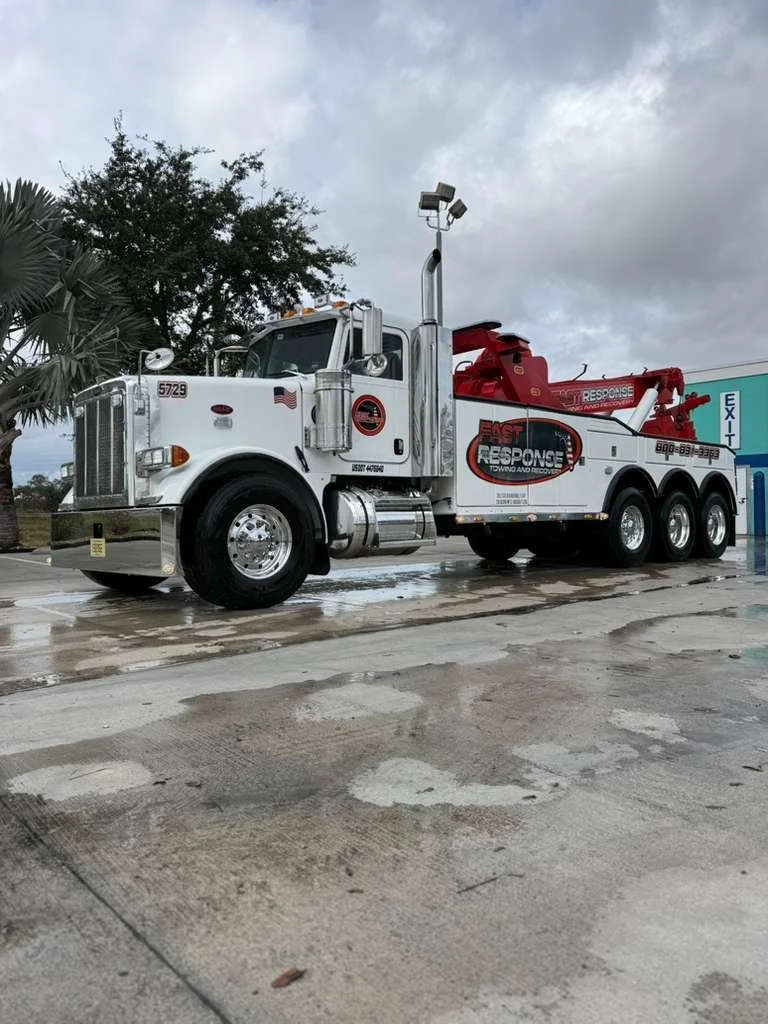 White tow truck with red and black branding parked on a wet pavement outside, with trees and a cloudy sky in the background.