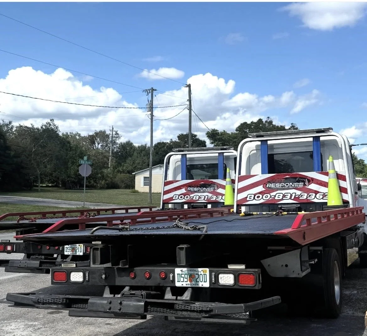 A tow truck parked on the side of the road with two service trucks on its flatbed, yellow safety cones, and Florida license plates, under a partly cloudy sky.